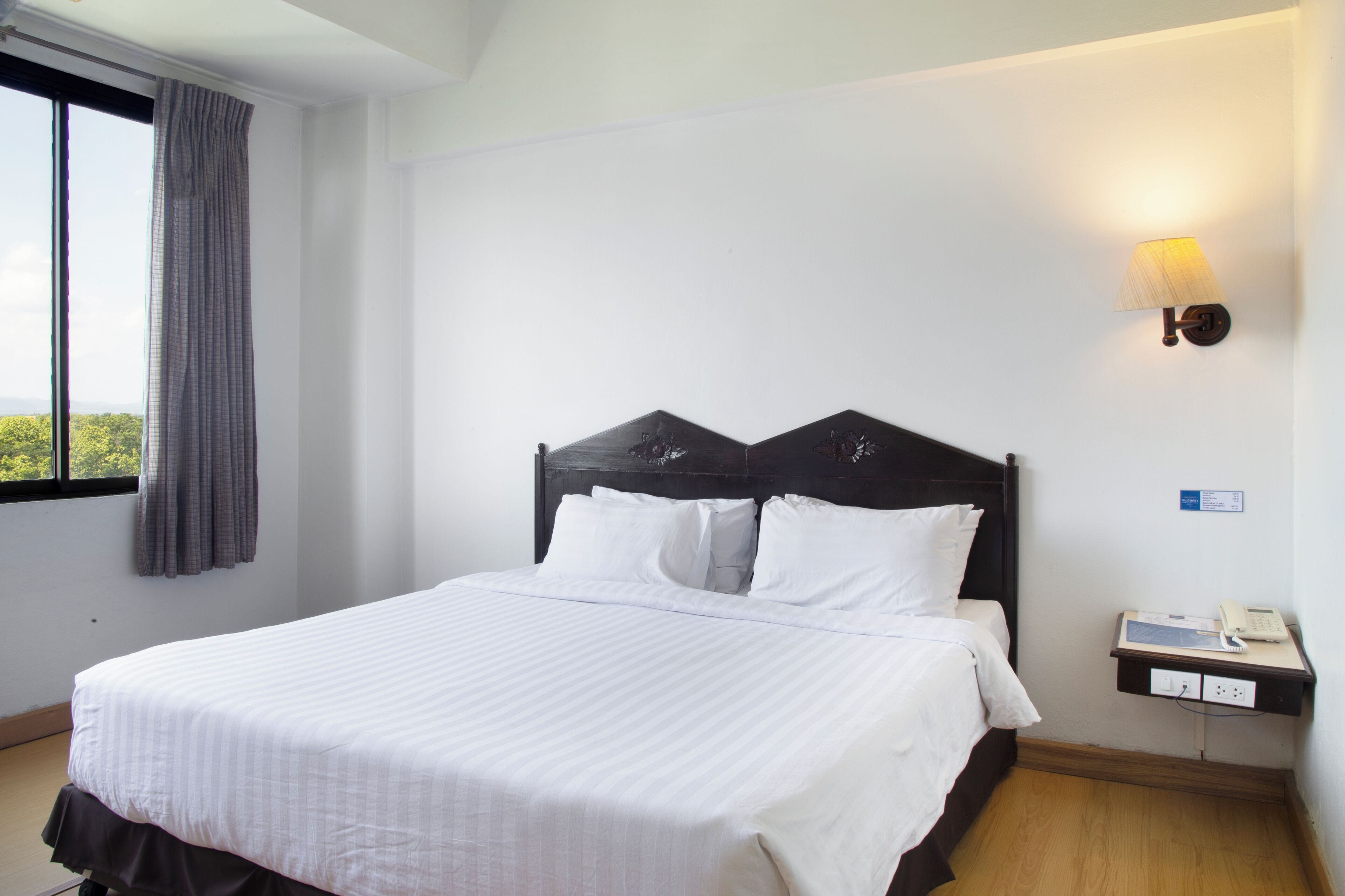 A hotel room with a bed, a nightstand with a phone, a window with curtains, and a wall-mounted light.