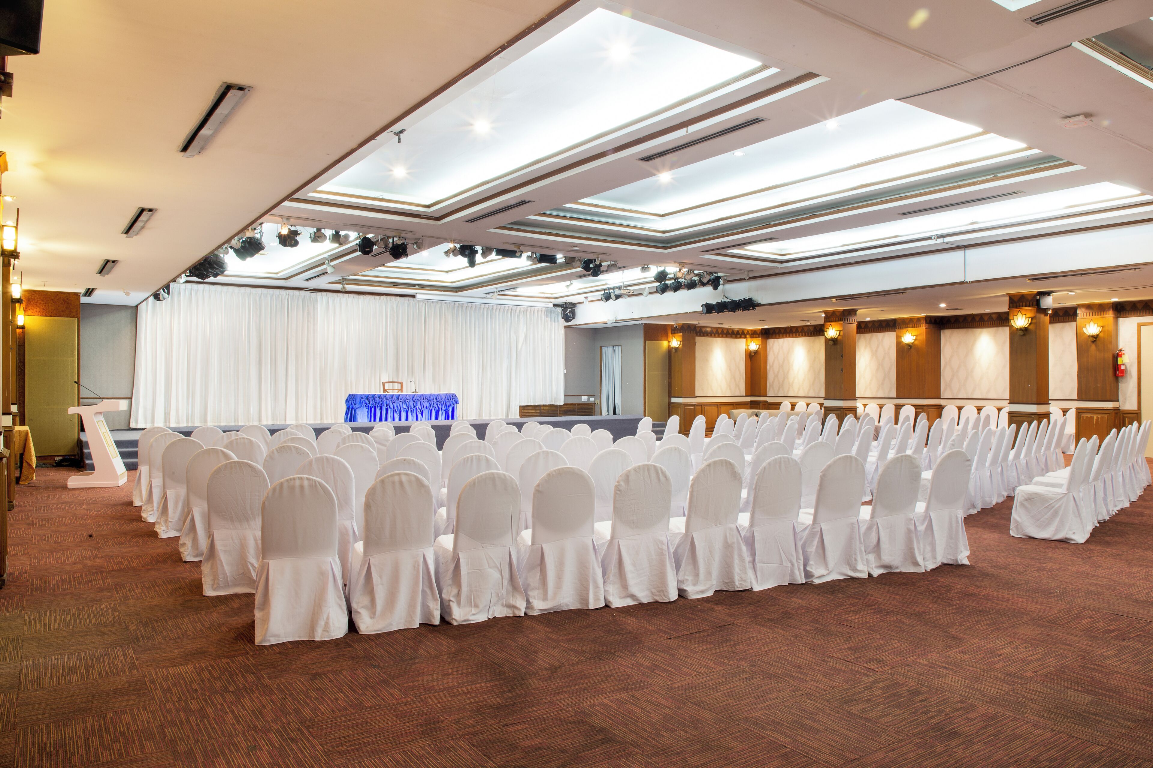 A conference room with rows of white chairs, a stage with a blue tablecloth, and a ceiling with skylights.