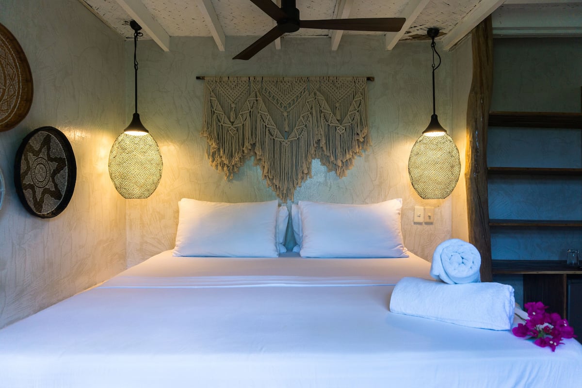 A neatly made bed with white linens, a macrame wall hanging, and two hanging lamps.