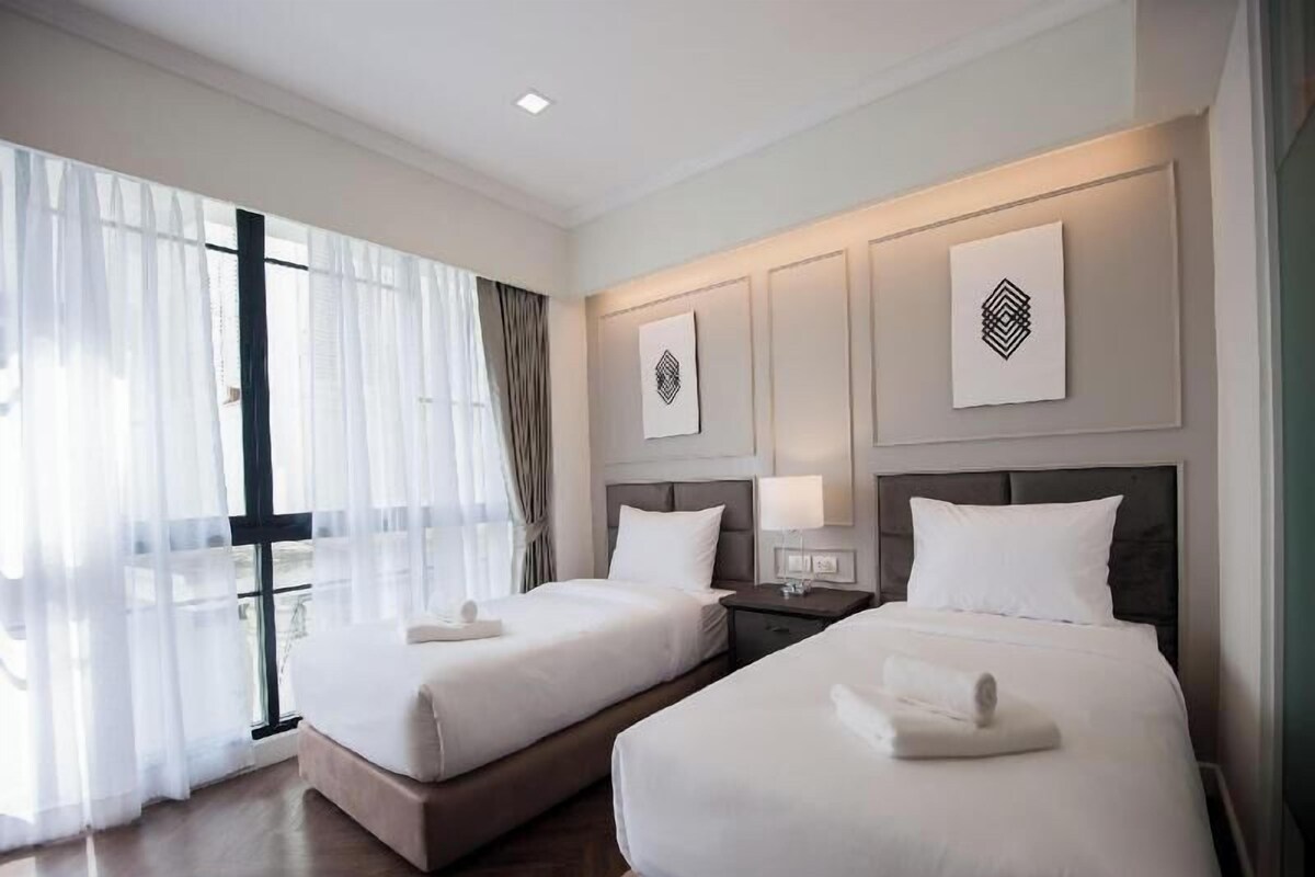 A hotel room with two beds, a large window with sheer curtains, and artwork on the walls.