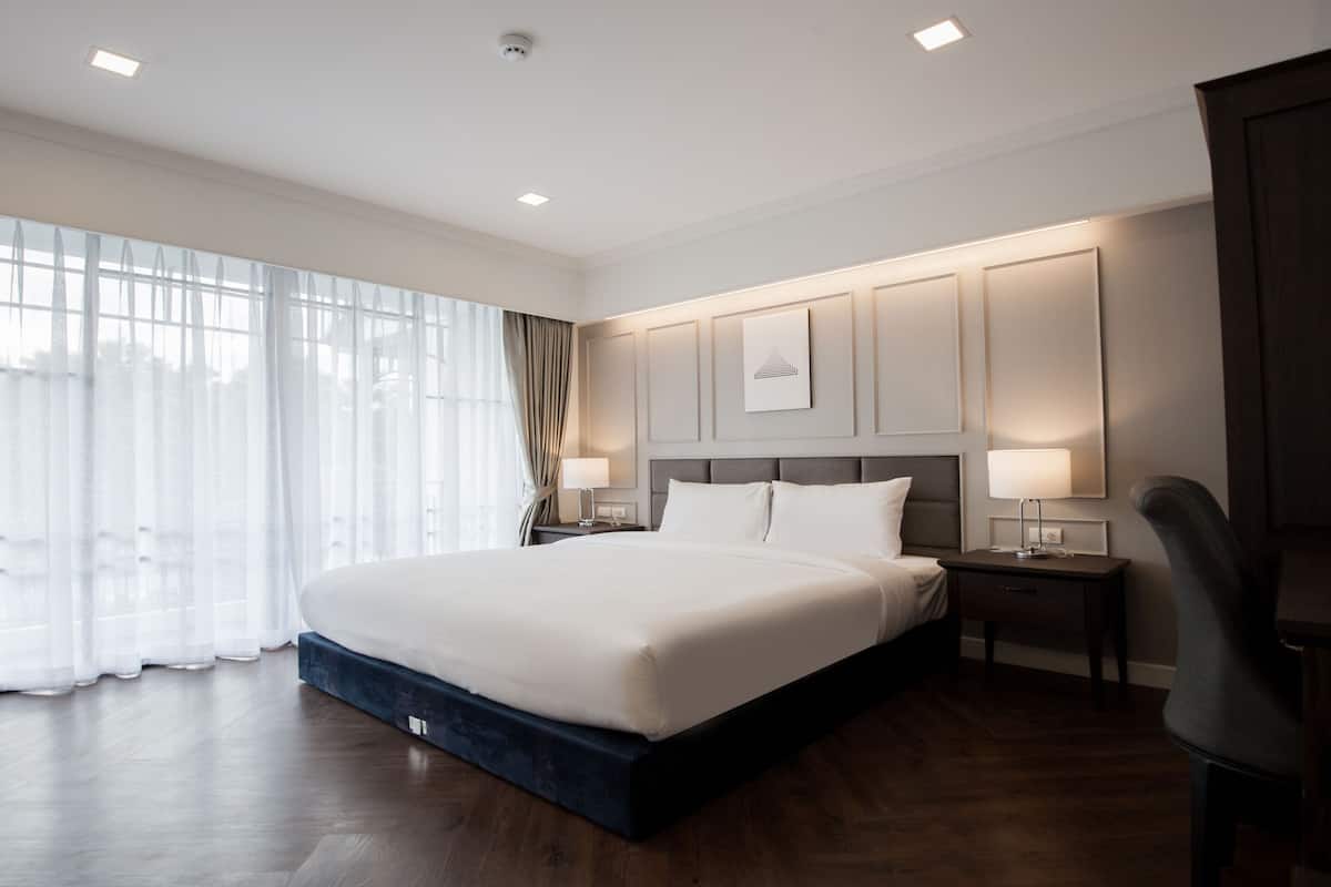 Habitación de hotel con una cama grande, dos mesitas de noche, una silla y un ventanal con cortinas de encaje.