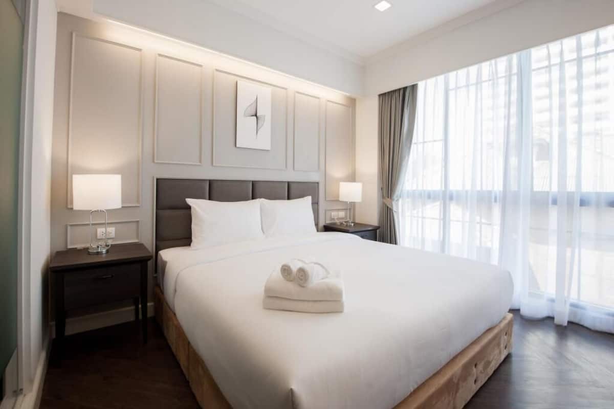 Habitación de hotel con una cama grande, dos mesitas de noche con lámparas, un cabecero con un reloj y una ventana con cortinas.