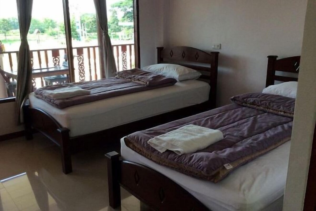 Een kamer met twee houten bedden, elk met hoofdbord en kussen. Er is een raam met gordijnen, en een balkon is te zien buiten.