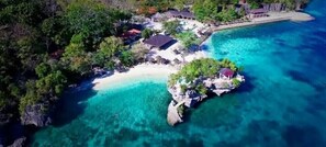 Vista aérea de um resort em ilha tropical com águas azul-turquesa cristalinas, uma praia arenosa e várias estruturas, incluindo um prédio principal e algumas cabanas.