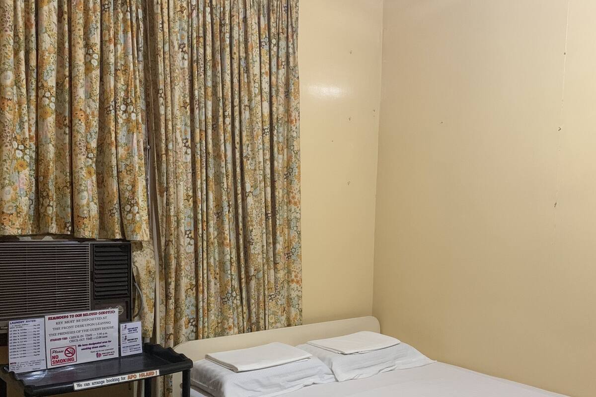 Una cama individual con ropa de cama blanca, una mesita de noche de madera con una bandeja negra y una cortina floral.