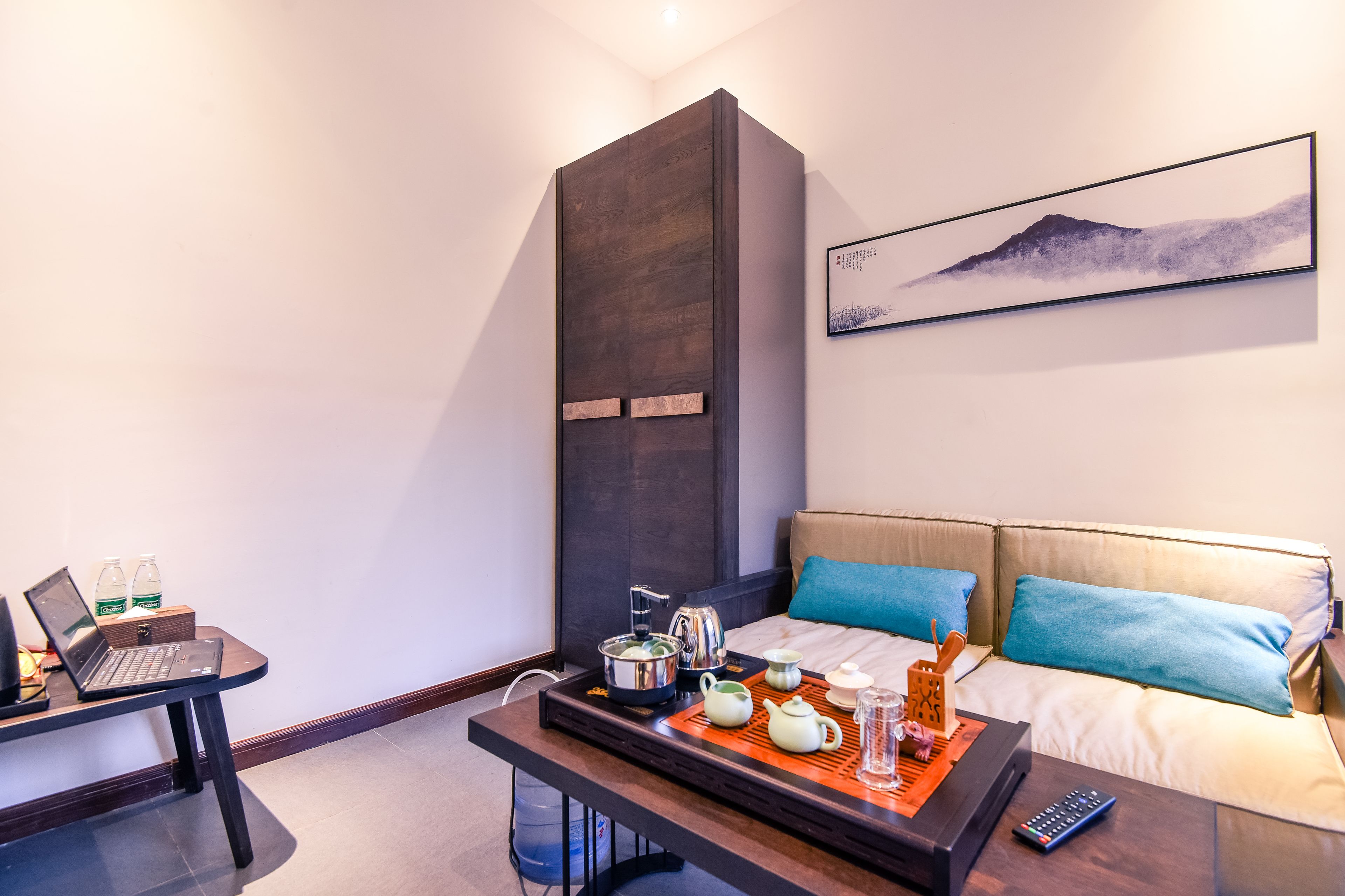 A modern hotel room with a bed, a desk with a laptop, a tray with tea and cups, and a painting on the wall.