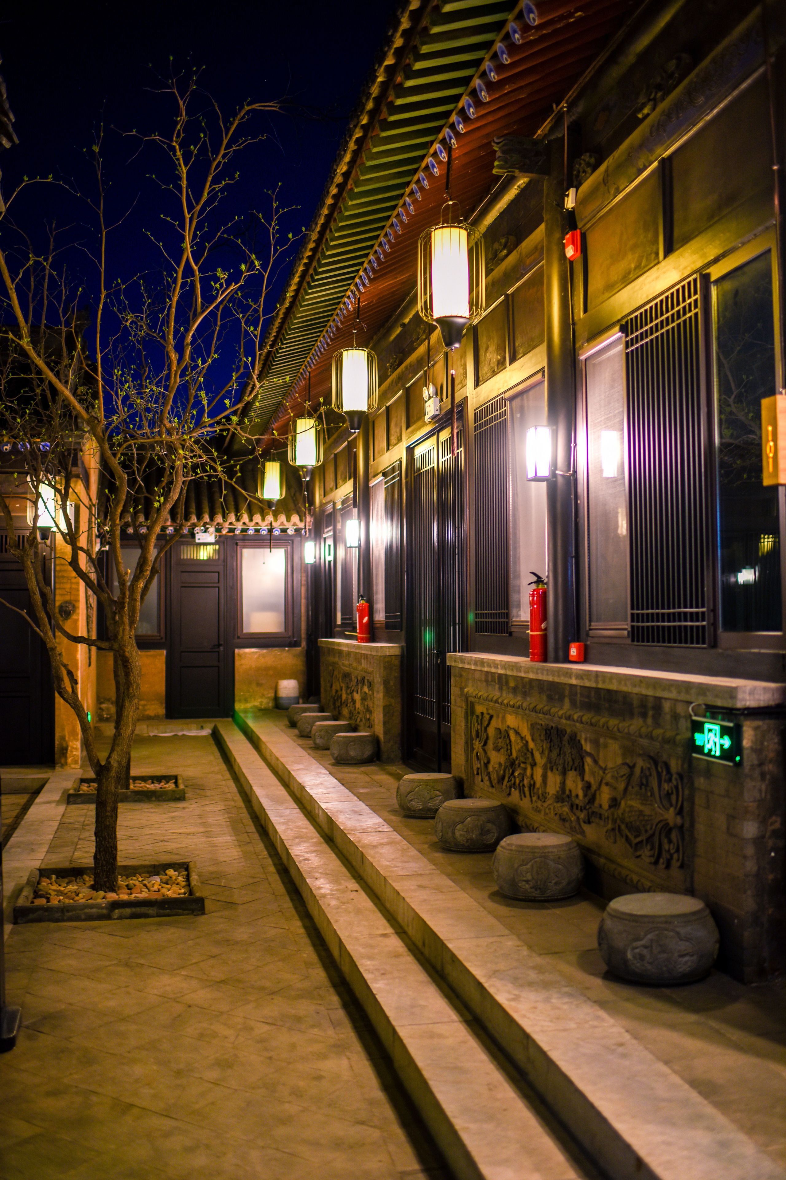 A traditional courtyard with stone lanterns, a tree, and a building with a tiled roof and wooden doors.
