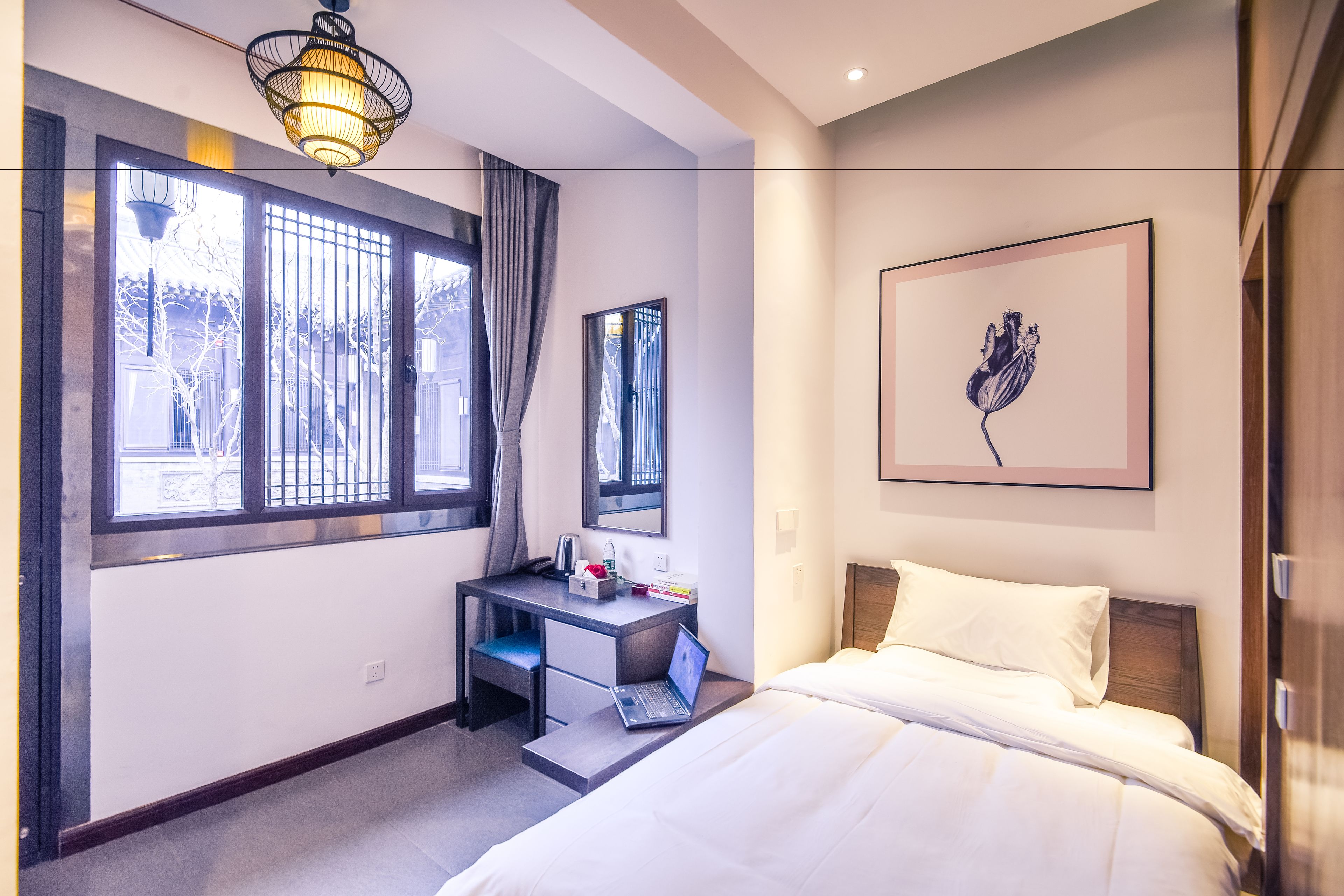 A modern hotel room with a bed, a desk with a laptop, a large window with curtains, and a framed artwork on the wall.