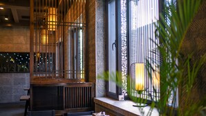 Interior de restaurante com mobiliário de madeira, uma janela com grades metálicas decorativas e uma planta em primeiro plano.