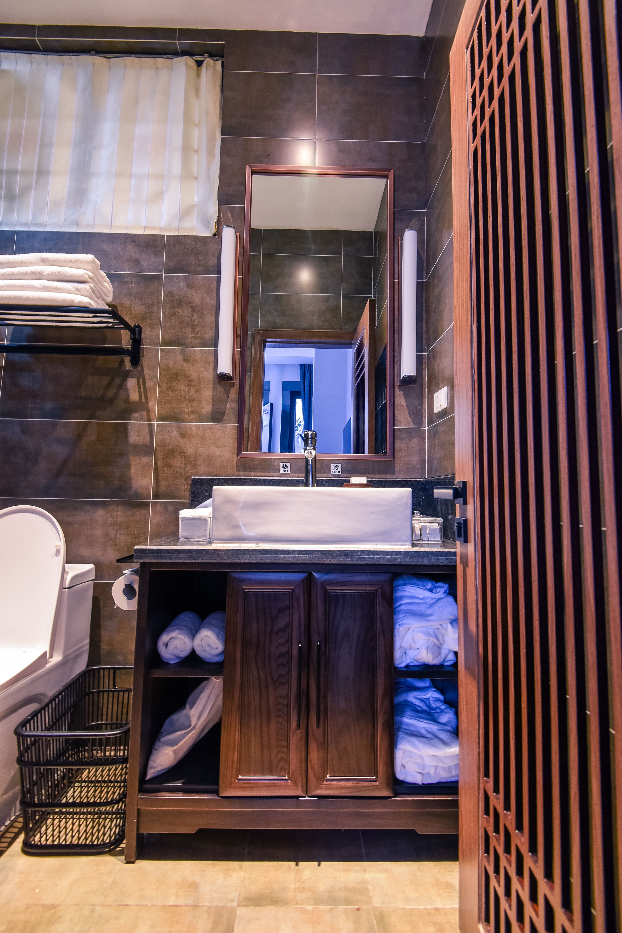A modern bathroom with a dark tiled wall, a wooden vanity with a sink, a mirror, and a towel rack.