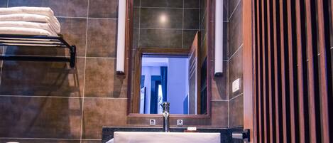 A modern bathroom with a dark tiled wall, a wooden vanity with a sink, a mirror, and a towel rack.