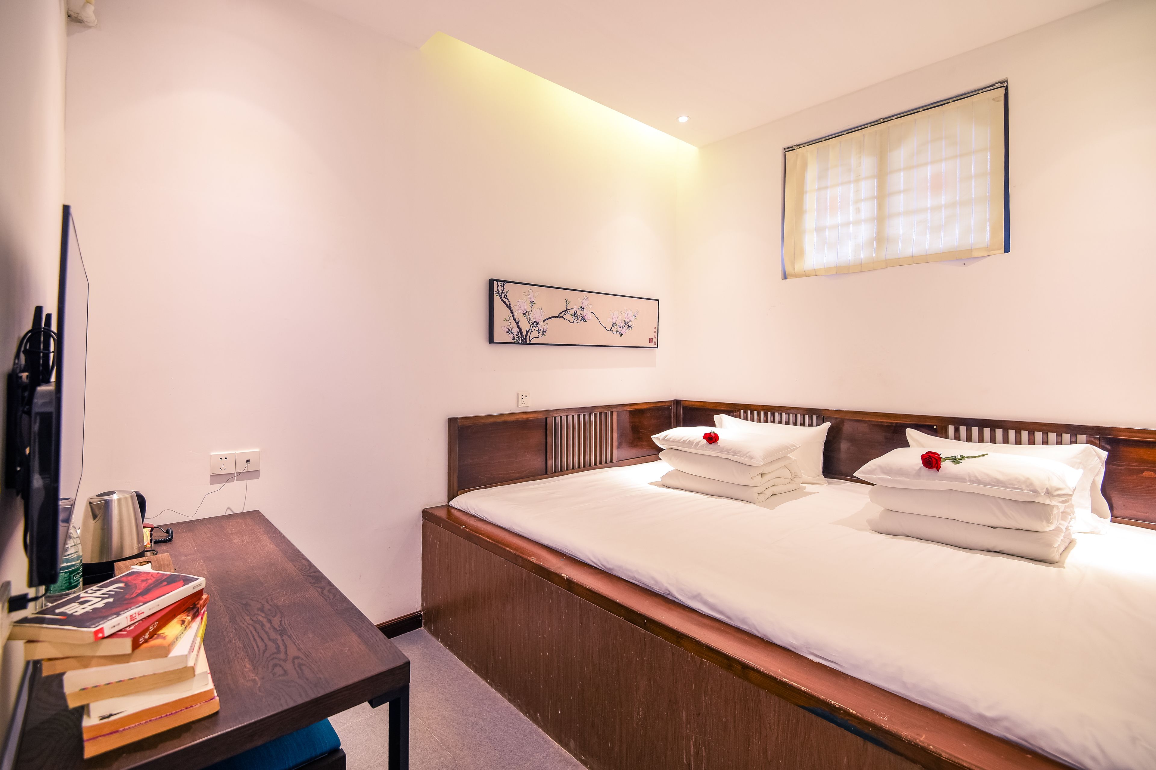 A neatly made bed with white linens and pillows, a wooden bedside table with books and a kettle, a framed artwork on the wall, and a window with blinds.