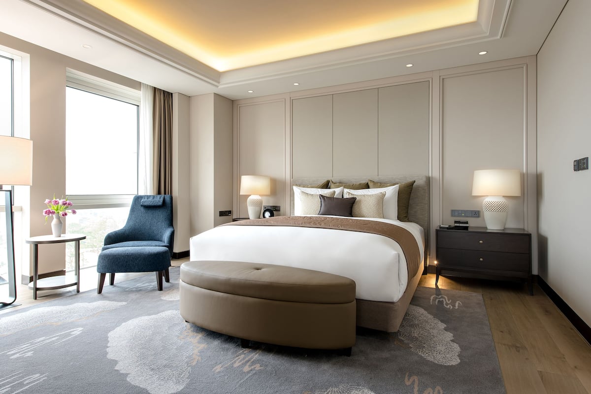 A modern hotel room with a large bed, two bedside tables with lamps, a blue armchair, a small round table with a vase of flowers, and a large window with curtains.