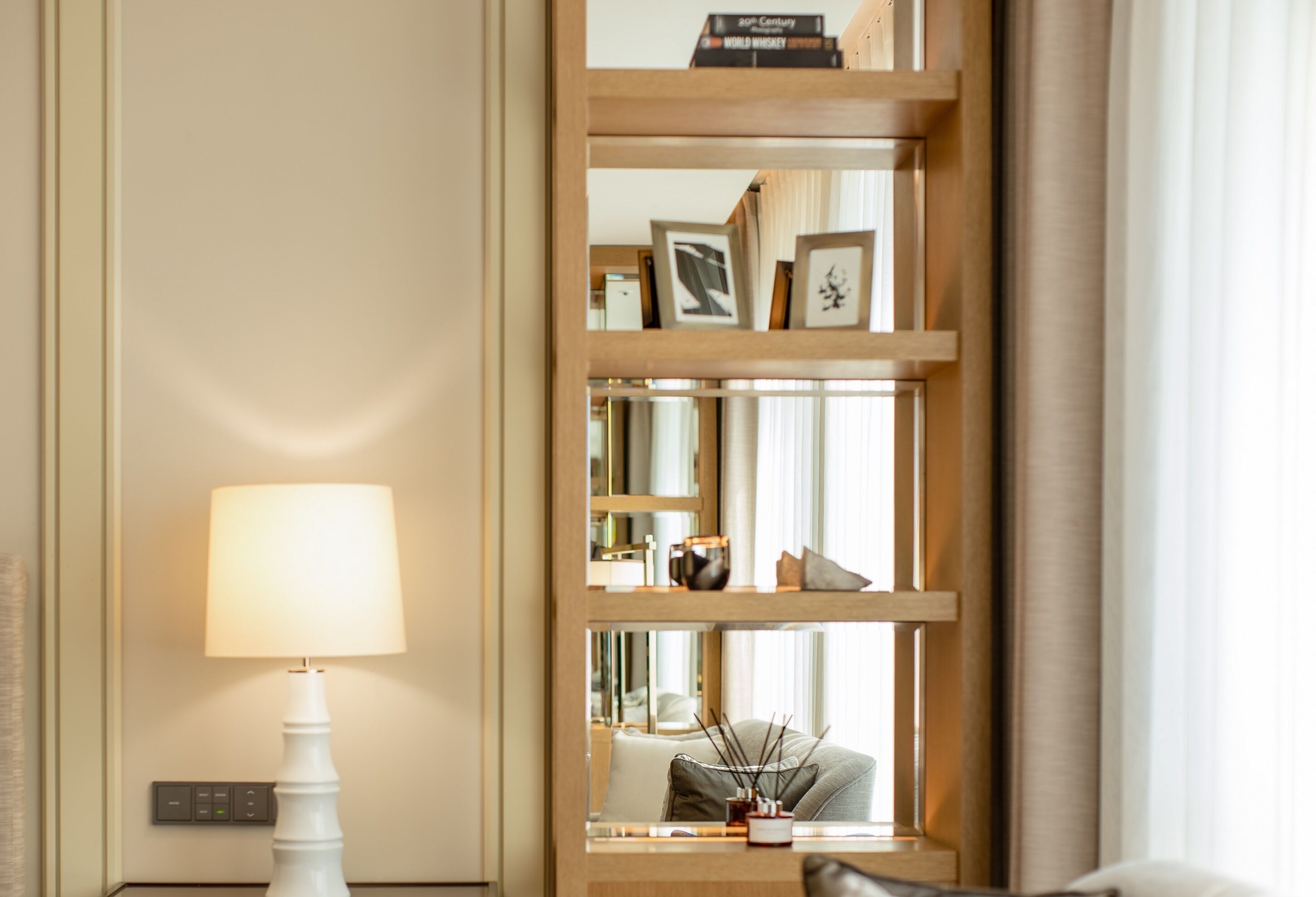 Una habitación de hotel con una lámpara blanca, una estantería de madera con marcos de fotos y un espejo que refleja la habitación.