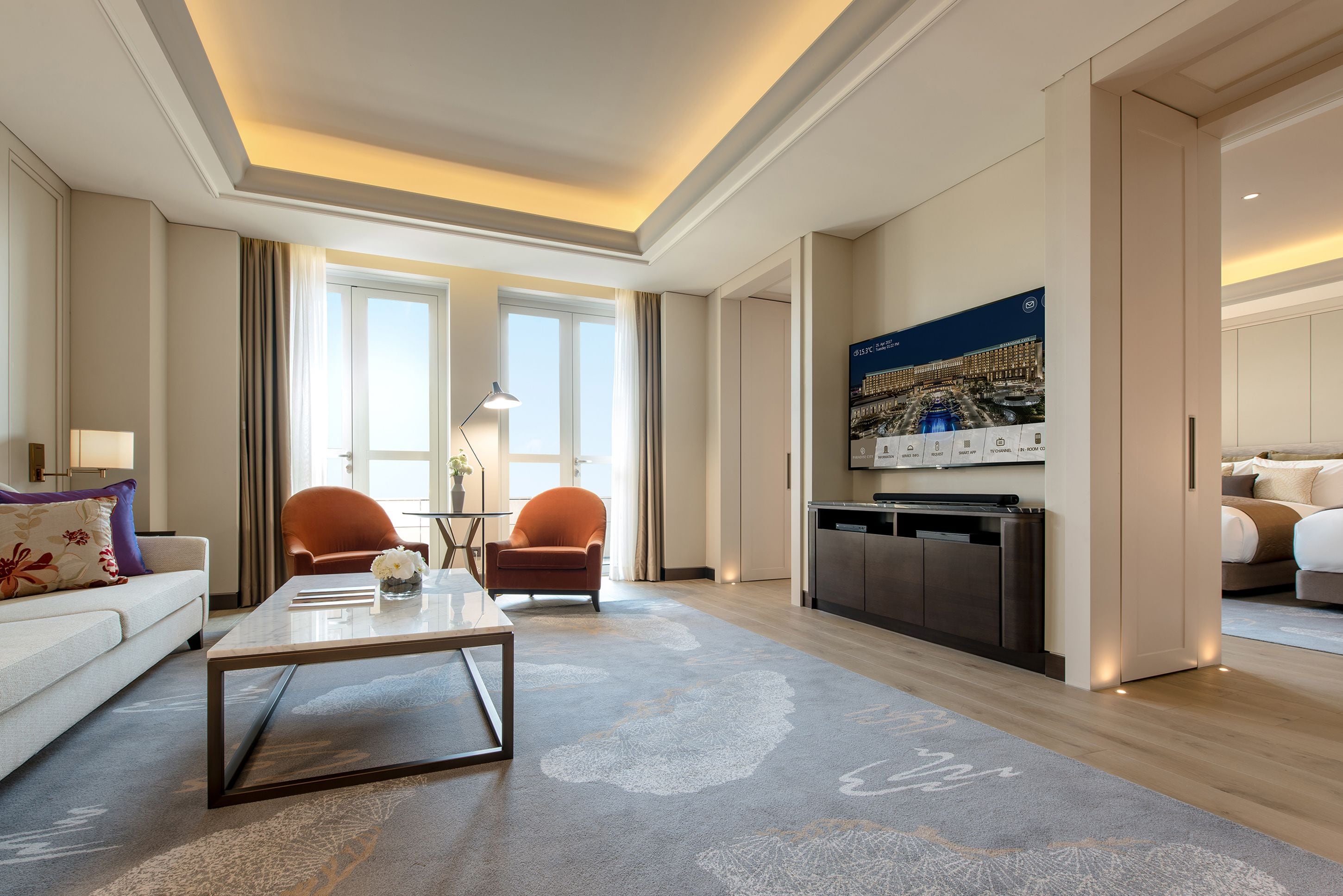 A modern hotel room with a sofa, armchairs, a coffee table, a dining table, a kitchen area, and a large cityscape painting on the wall.