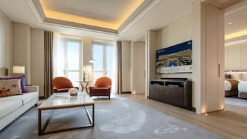 A modern hotel room with a sofa, armchairs, a coffee table, a dining table, a kitchen area, and a large cityscape painting on the wall.