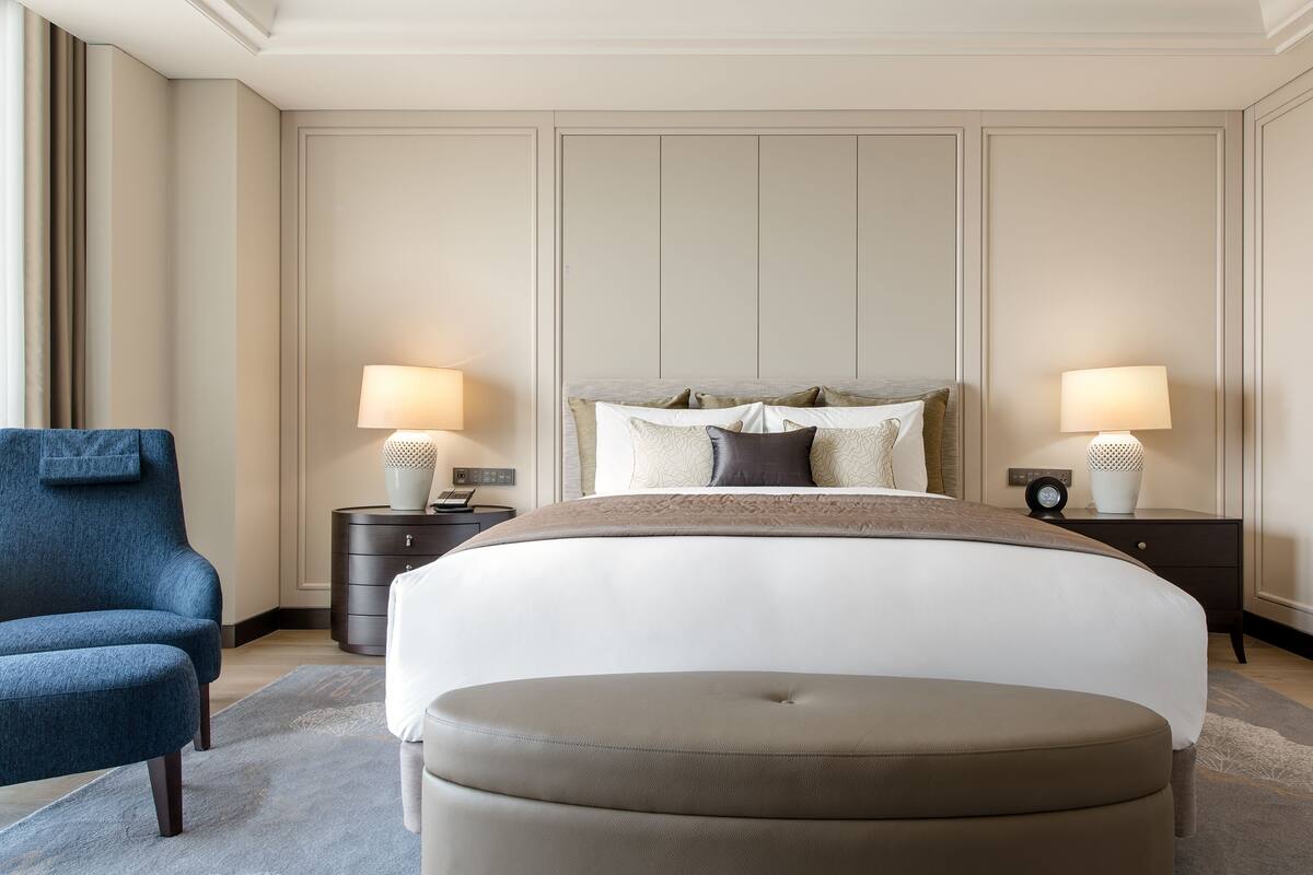 A hotel room with a large bed, two bedside tables with lamps, a blue armchair, and a round ottoman.