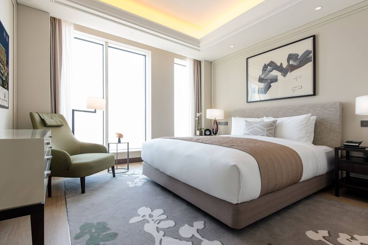 A modern hotel room with a large bed, a green armchair, a desk, and a view of the city through large windows.
