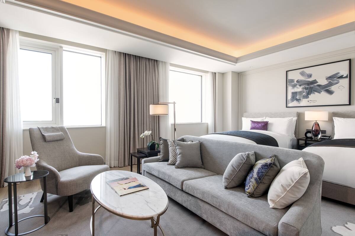A hotel room with a grey sofa, a small round table, a bed with a black headboard, and a large window with curtains.