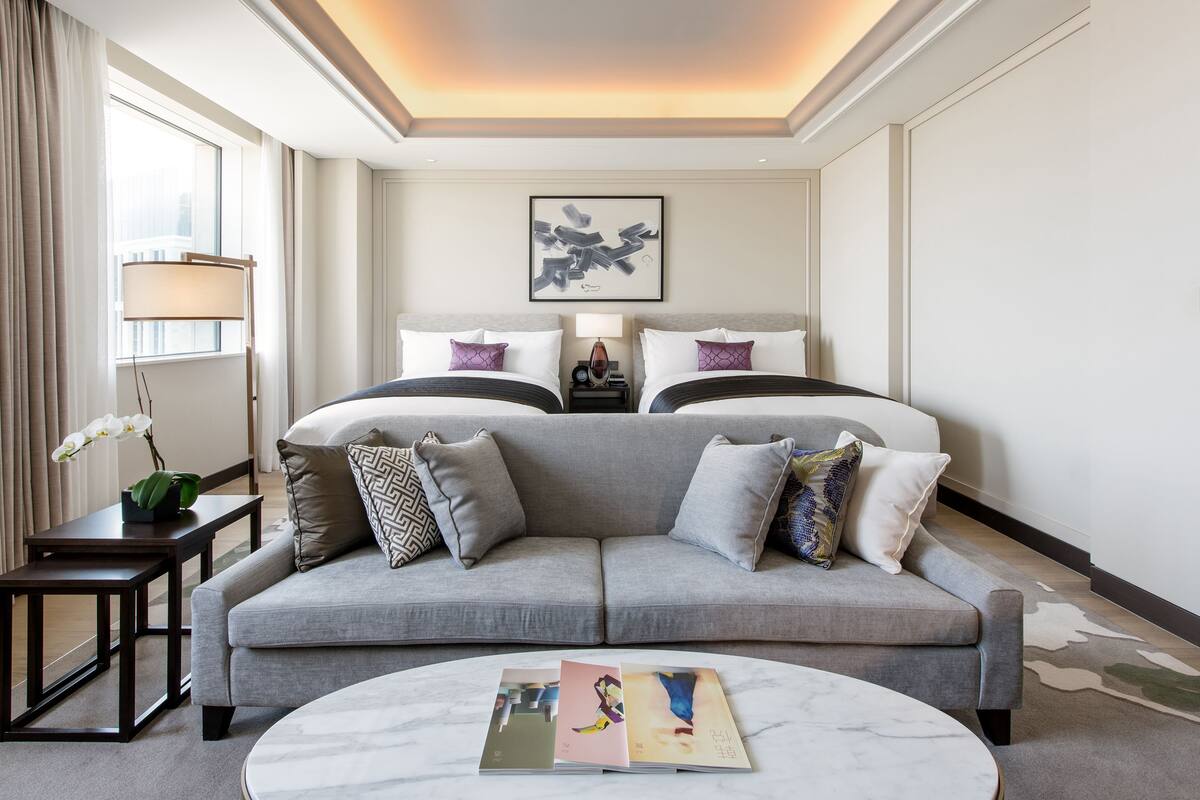 A modern hotel room with a grey sofa, two beds, a marble coffee table, and a framed abstract artwork on the wall.