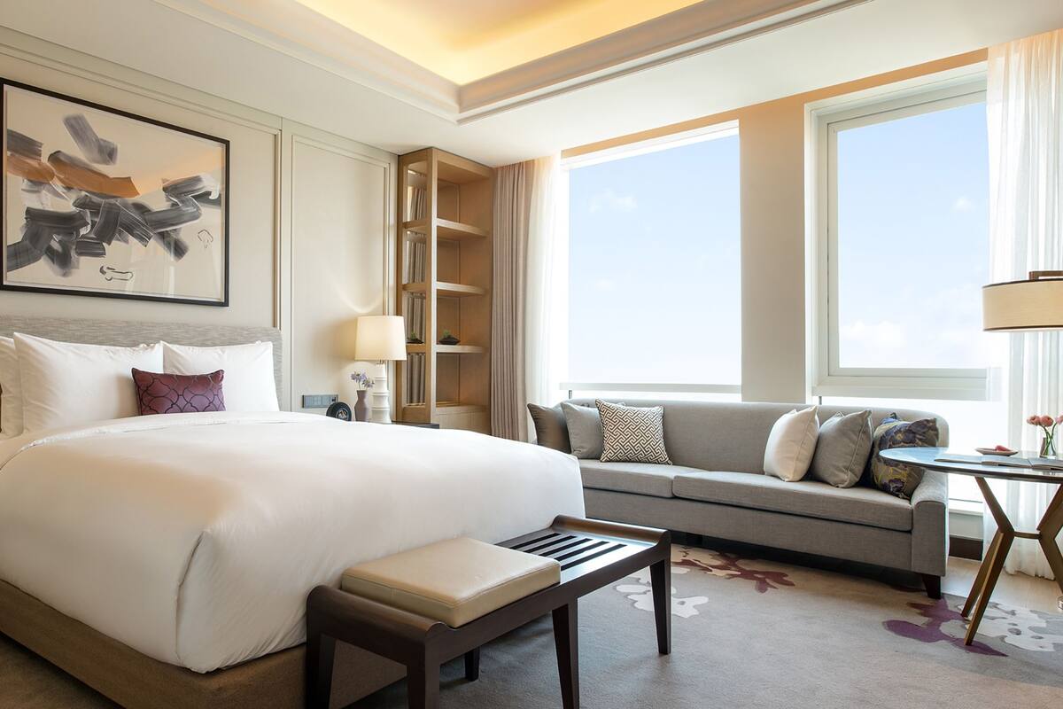 A hotel room with a large bed, a sofa, a small table, and a chair. There is a bookshelf and a window with curtains.