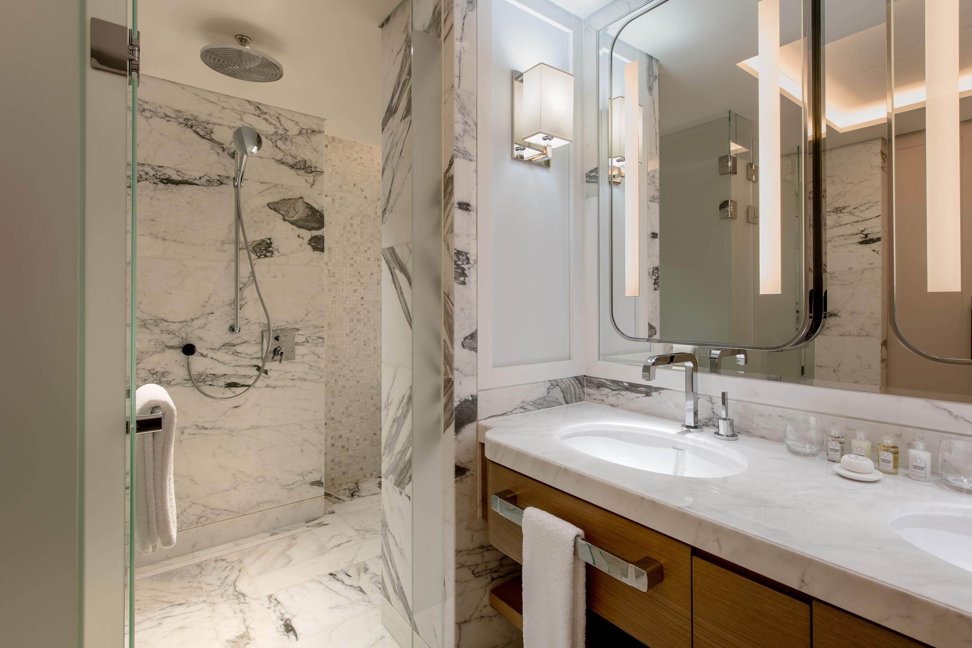 A modern bathroom with a glass shower enclosure, marble walls, a large mirror, and a vanity with two sinks.