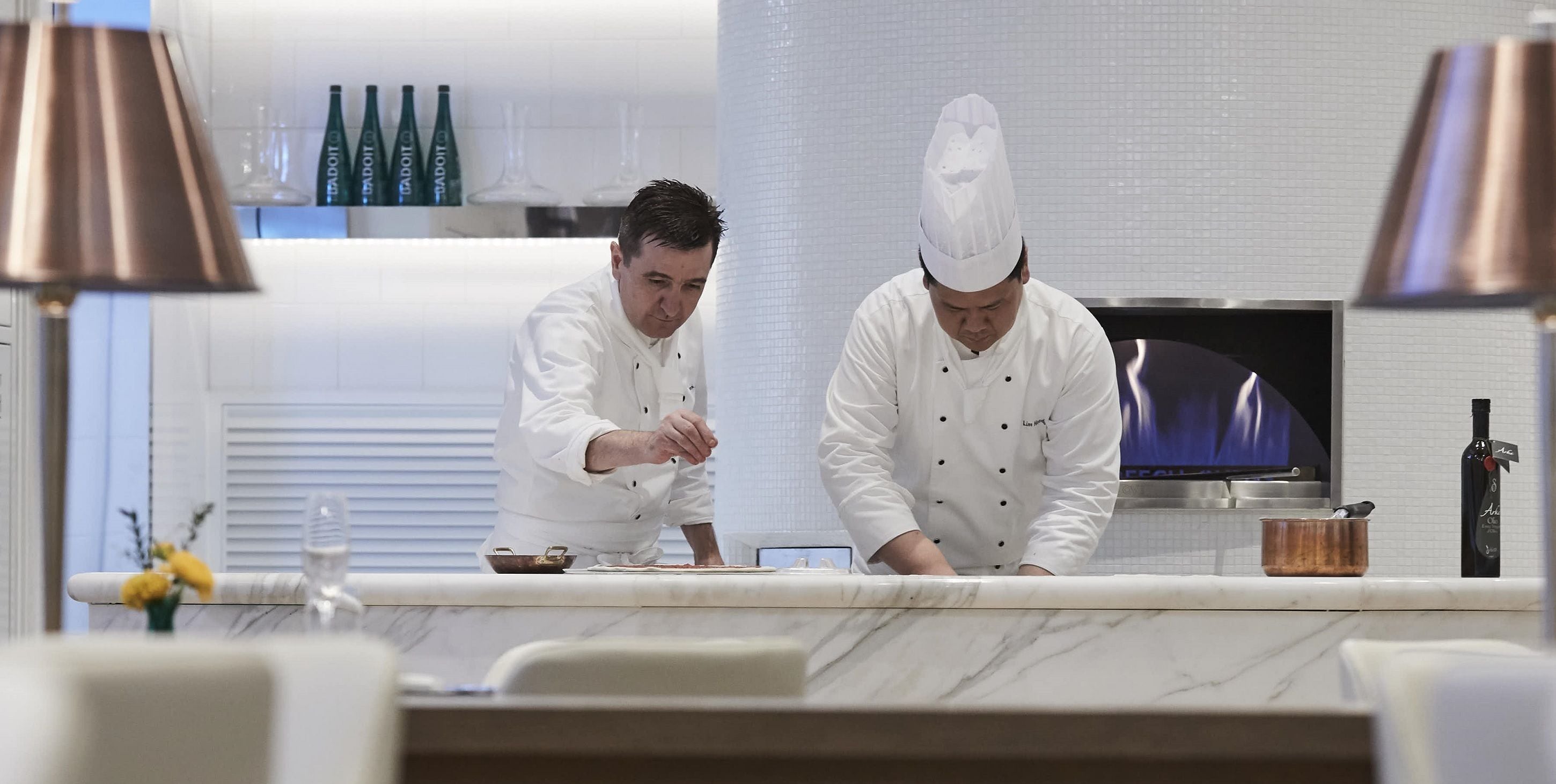 Two chefs in a modern kitchen with white cabinets, a marble countertop, and a television.