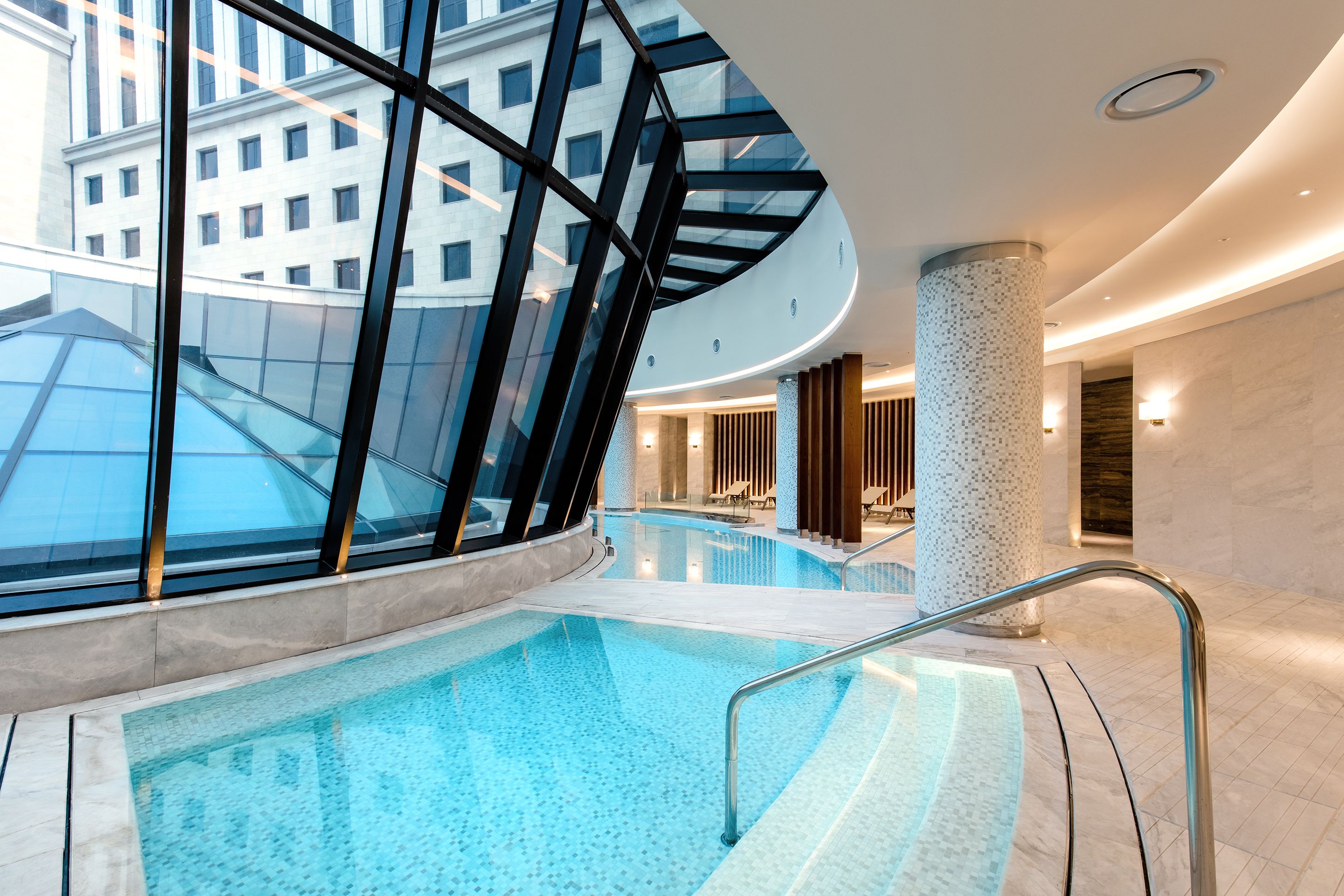 A modern indoor pool with glass walls, a curved ceiling, and a central column.