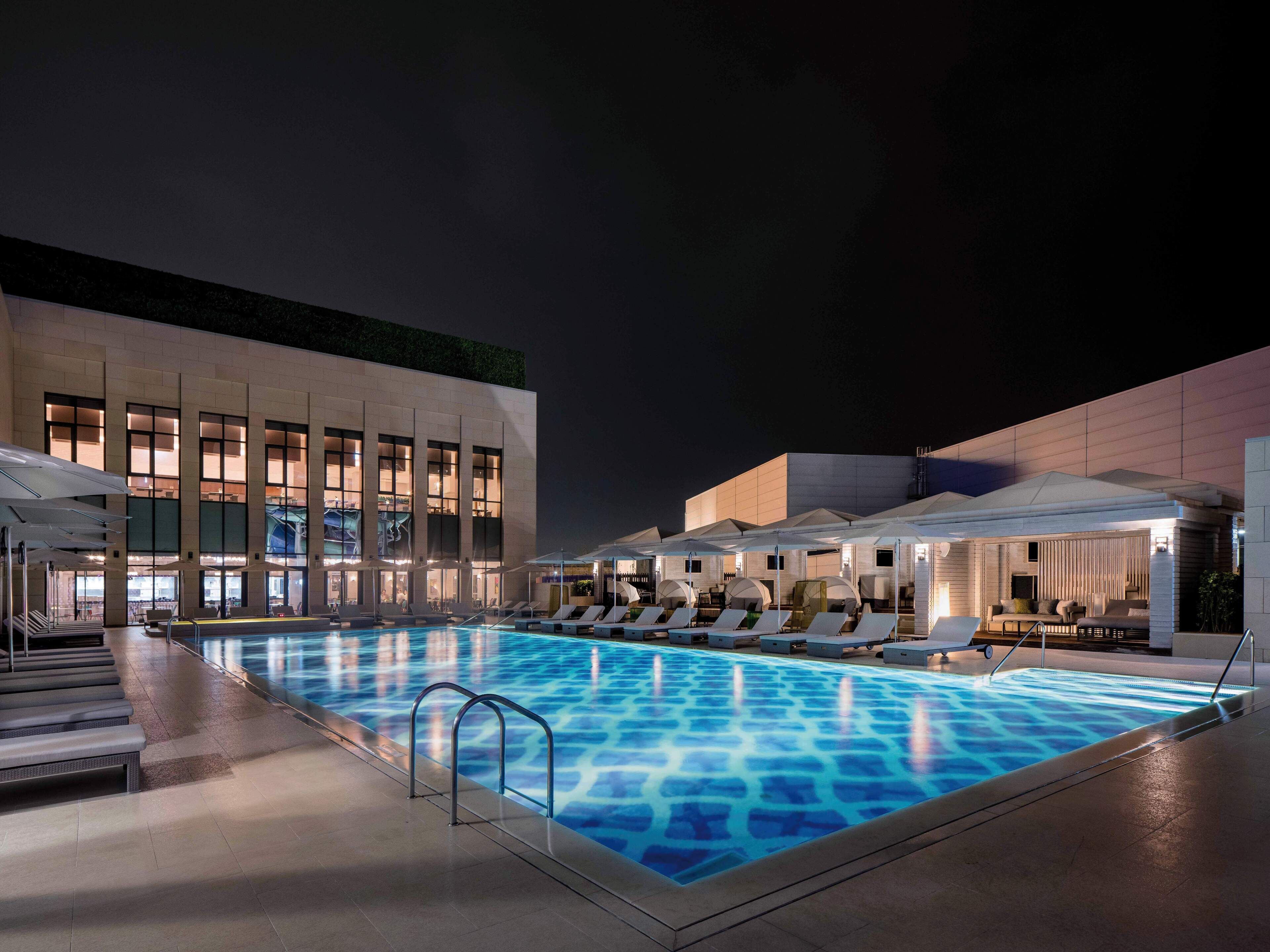 Un espace piscine nocturne doté de chaises longues et de parasols, un bâtiment aux grandes fenêtres et un design architectural moderne.