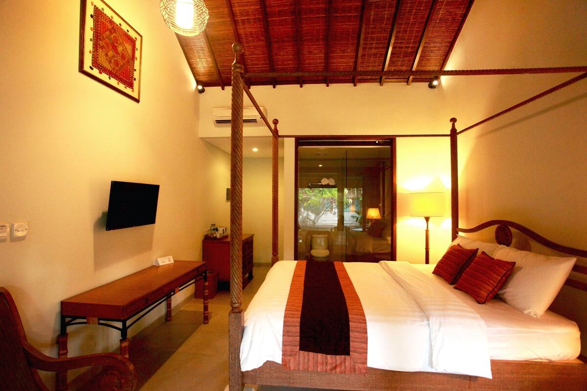 A hotel room with a four-poster bed, a flat-screen TV mounted on the wall, a wooden bench, and a desk.