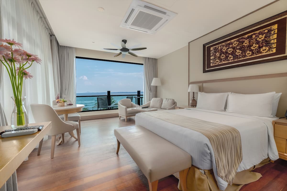 A hotel room with a large bed, a desk with a chair, a bench, a side table, a lamp, and a view of the sea.