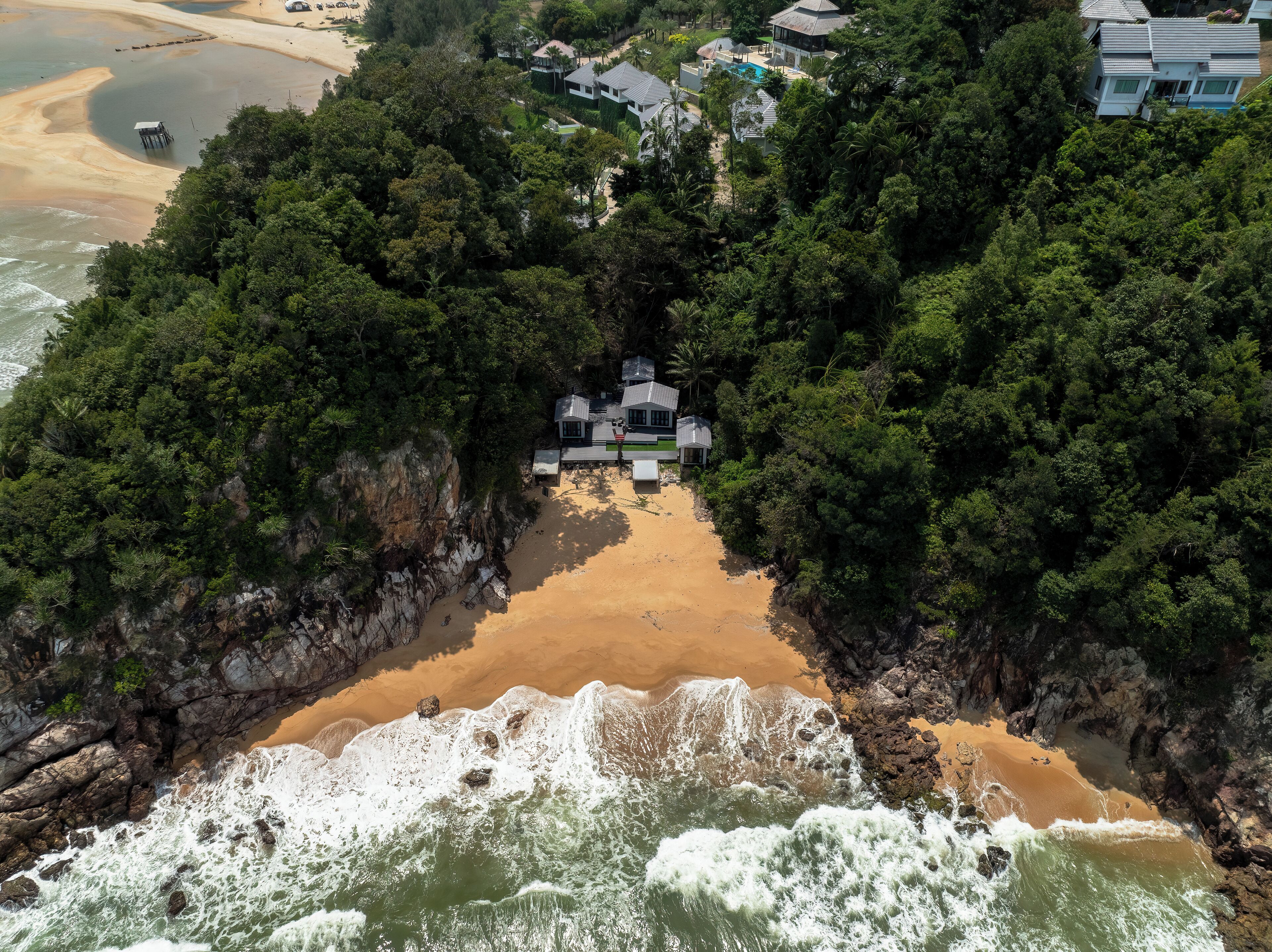 An aerial view of a secluded beach property surrounded by dense forest, featuring a central building with multiple rooms and a private pool.