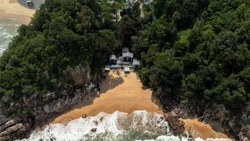 An aerial view of a secluded beach property surrounded by dense forest, featuring a central building with multiple rooms and a private pool.