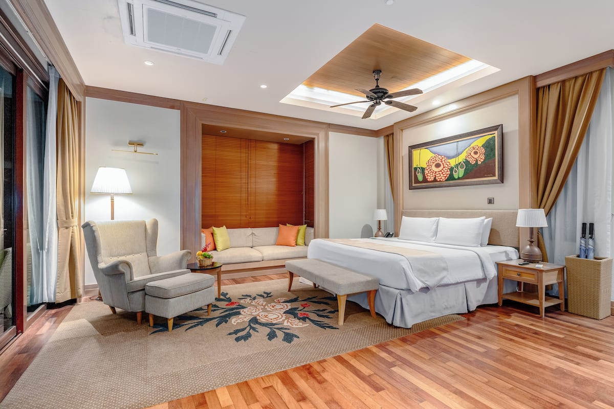 A hotel room with a bed, a sofa, a chair, a small table, a lamp, a ceiling fan, and a painting on the wall.