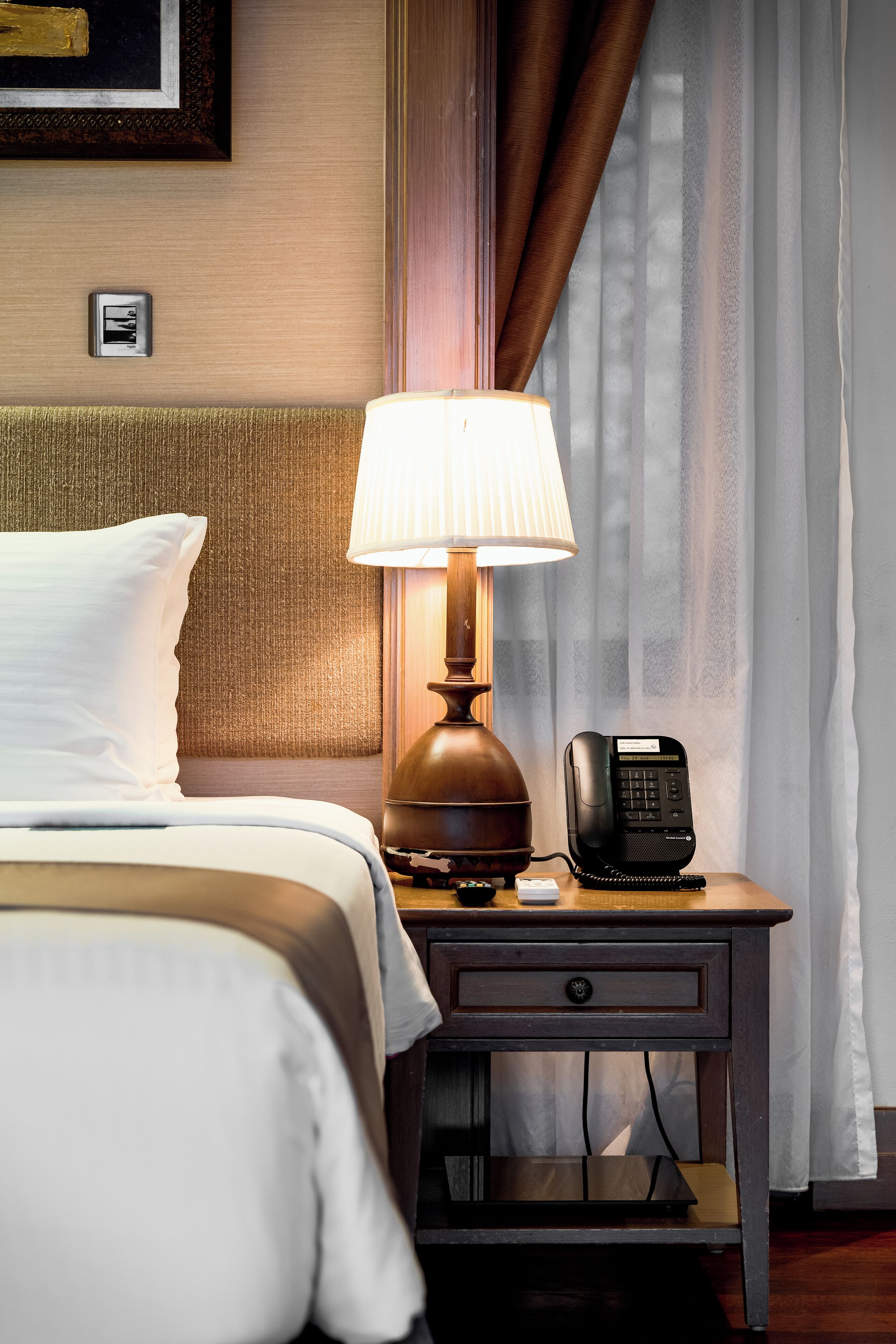 A neatly made bed with white linens, a wooden nightstand with a lamp, a telephone, and a small drawer.