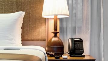 A neatly made bed with white linens, a wooden nightstand with a lamp, a telephone, and a small drawer.