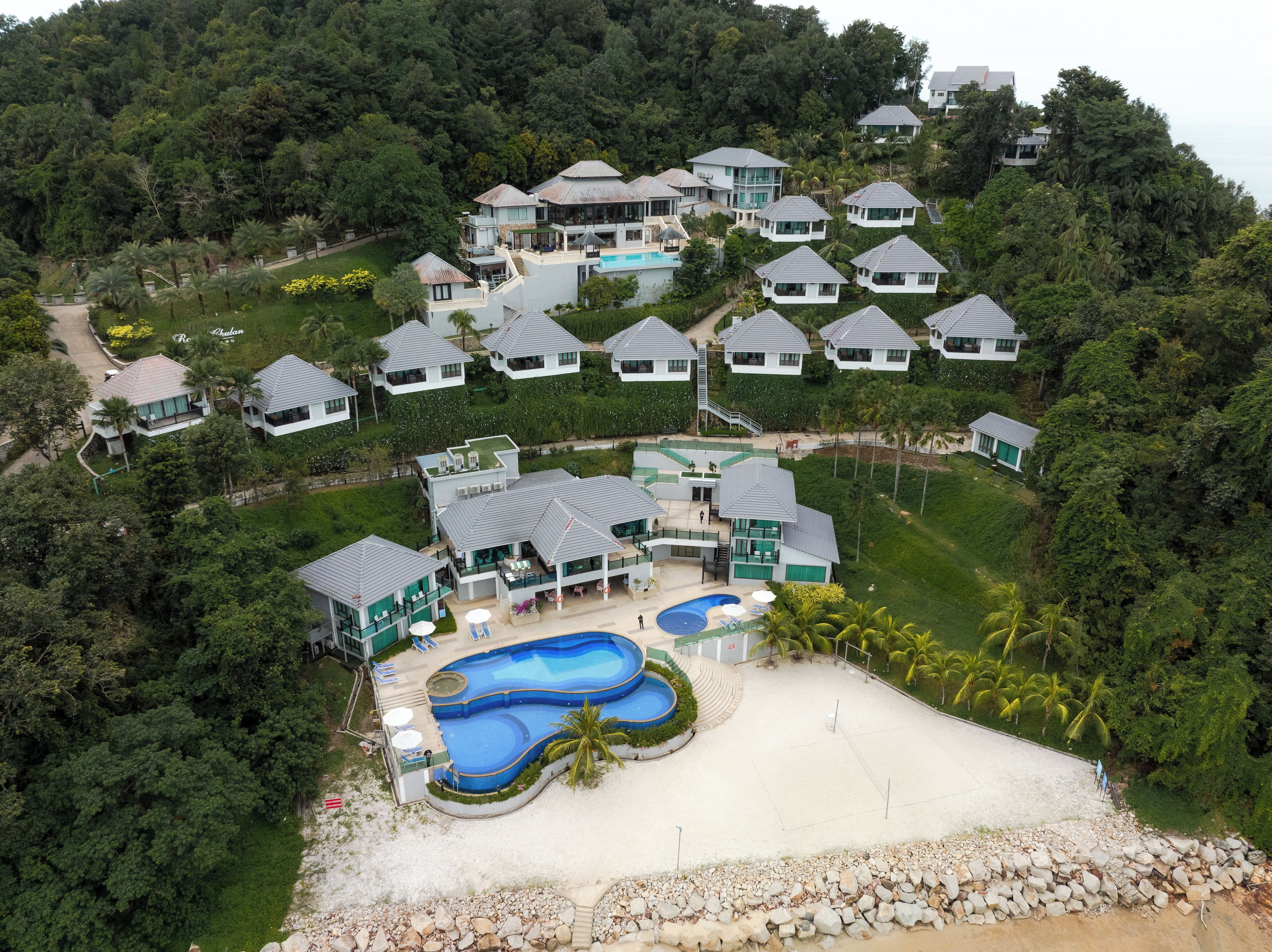 An aerial view of a resort with multiple villas, a central building with a pool, and a sandy beach area.