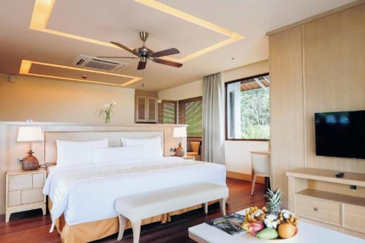 A hotel room with a bed, bedside tables, a flat-screen TV, a ceiling fan, and a window with blinds.