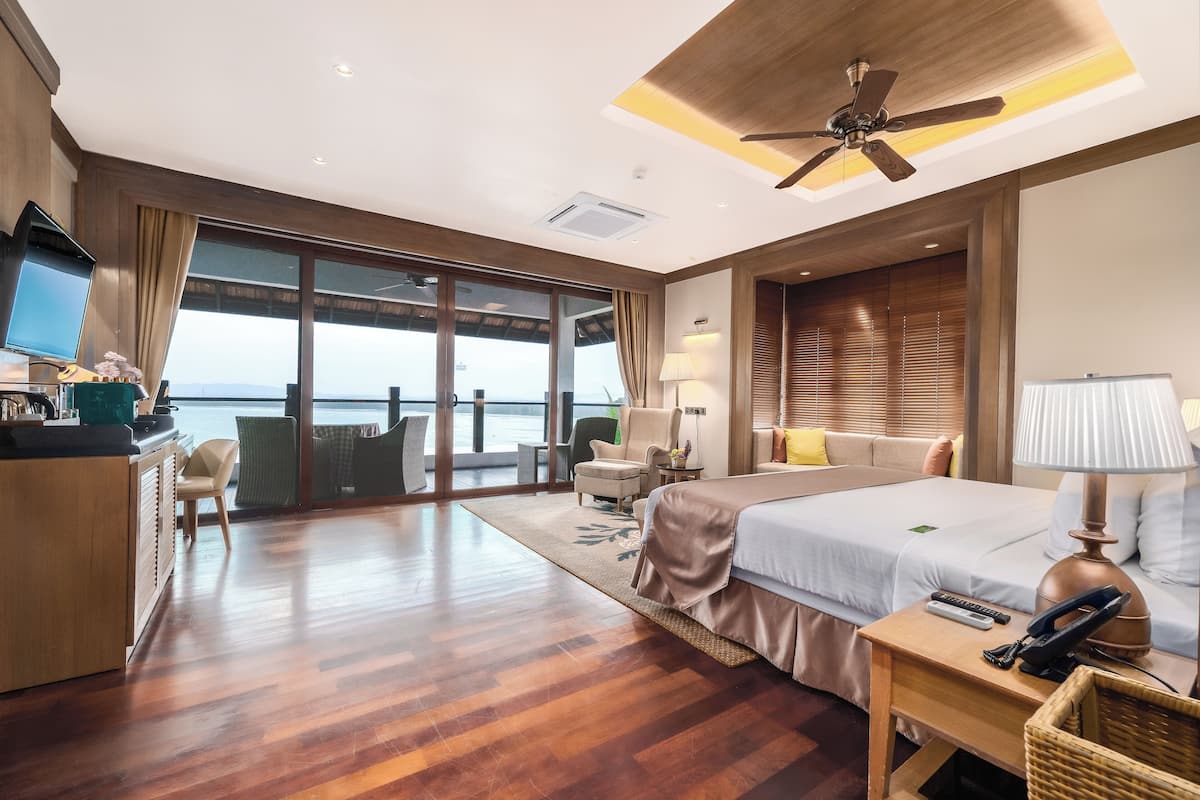 A hotel room with a large bed, a desk with a computer, a chair, a sofa, a TV, and a view of the sea.