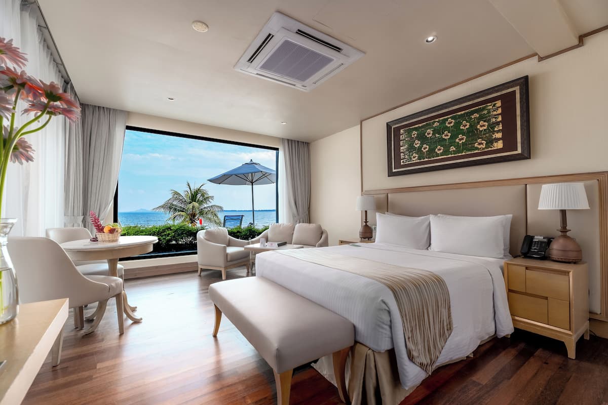 A hotel room with a large bed, a desk with a chair, a small table with a basket, a nightstand with a lamp, and a view of the ocean through a large window.
