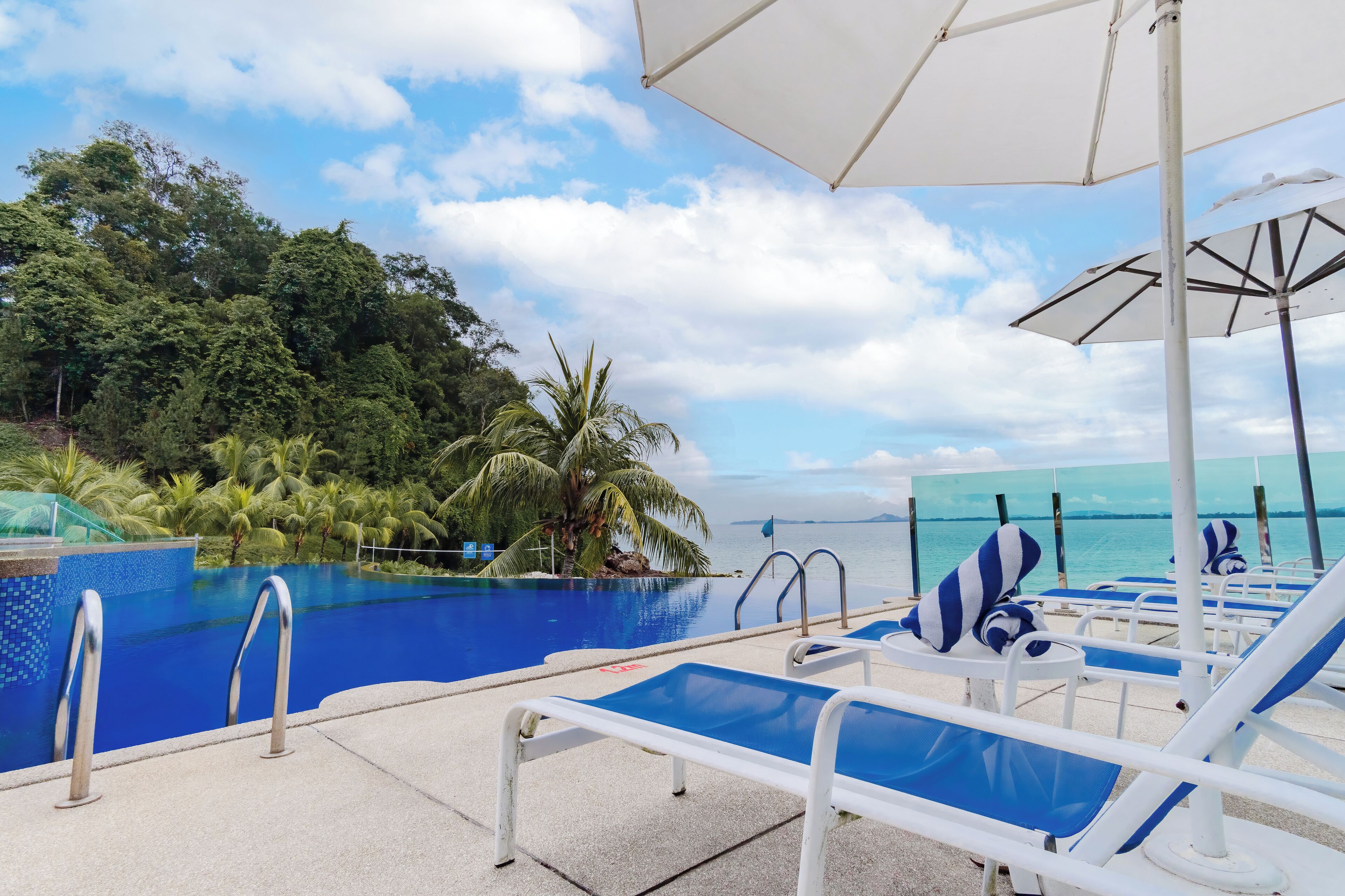 A poolside area with lounge chairs and umbrellas, a clear view of the ocean, and a tropical setting with palm trees.