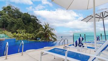 A poolside area with lounge chairs and umbrellas, a clear view of the ocean, and a tropical setting with palm trees.