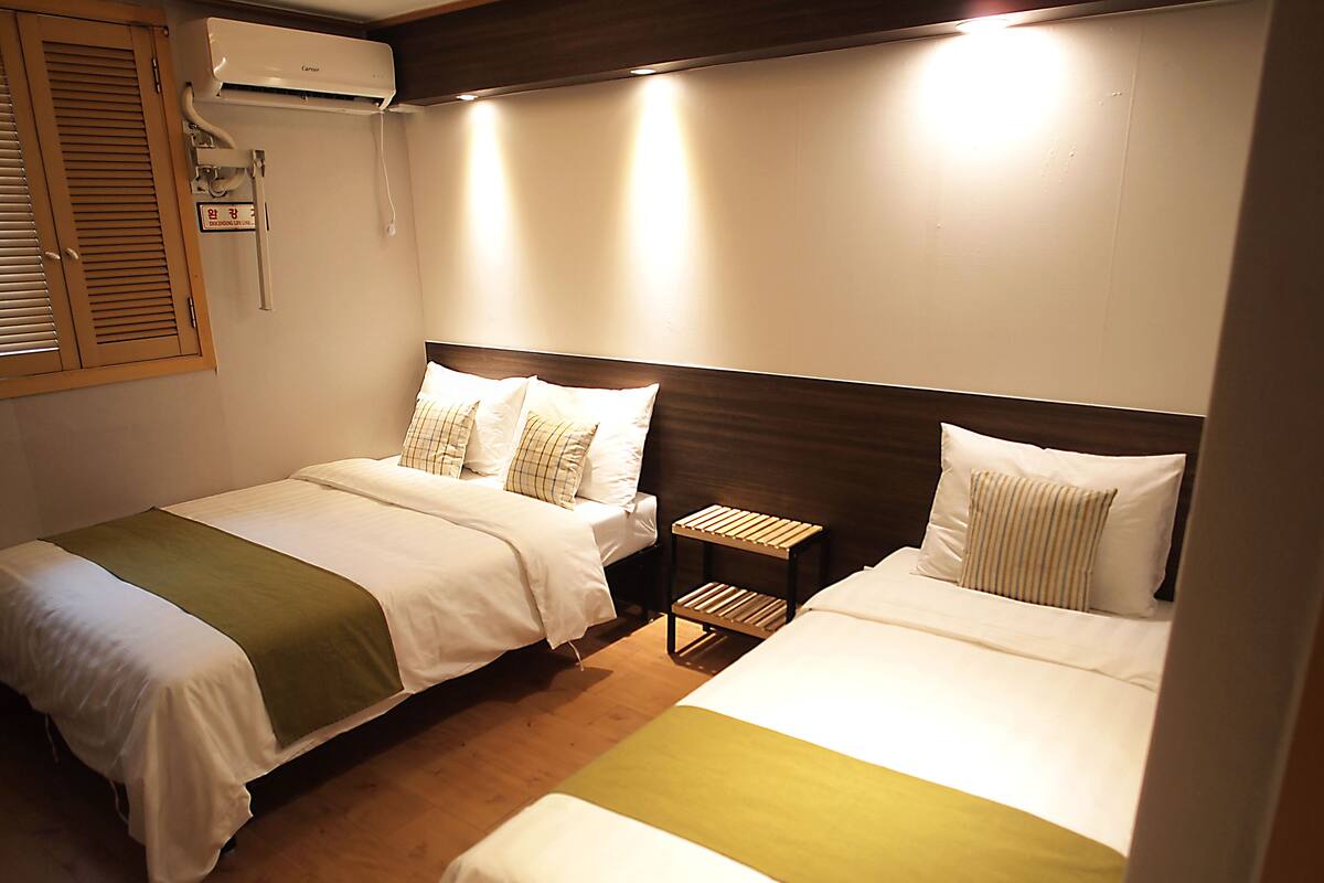 Habitación de hotel con dos camas individuales, cabecero de madera, mesita de noche pequeña y aire acondicionado de pared.
