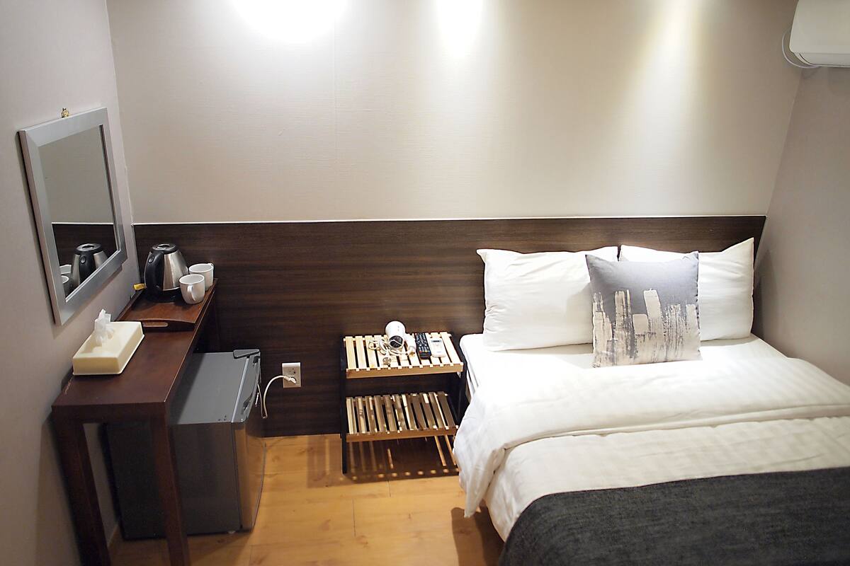 Una habitación de hotel con cama, mesita de noche, espejo y un pequeño rincón de descanso con una silla y una mesita.