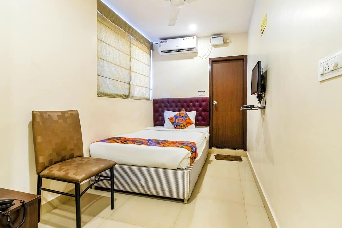 A small, neatly arranged bedroom with a single bed, a chair, an air conditioning unit, a television, and a door.