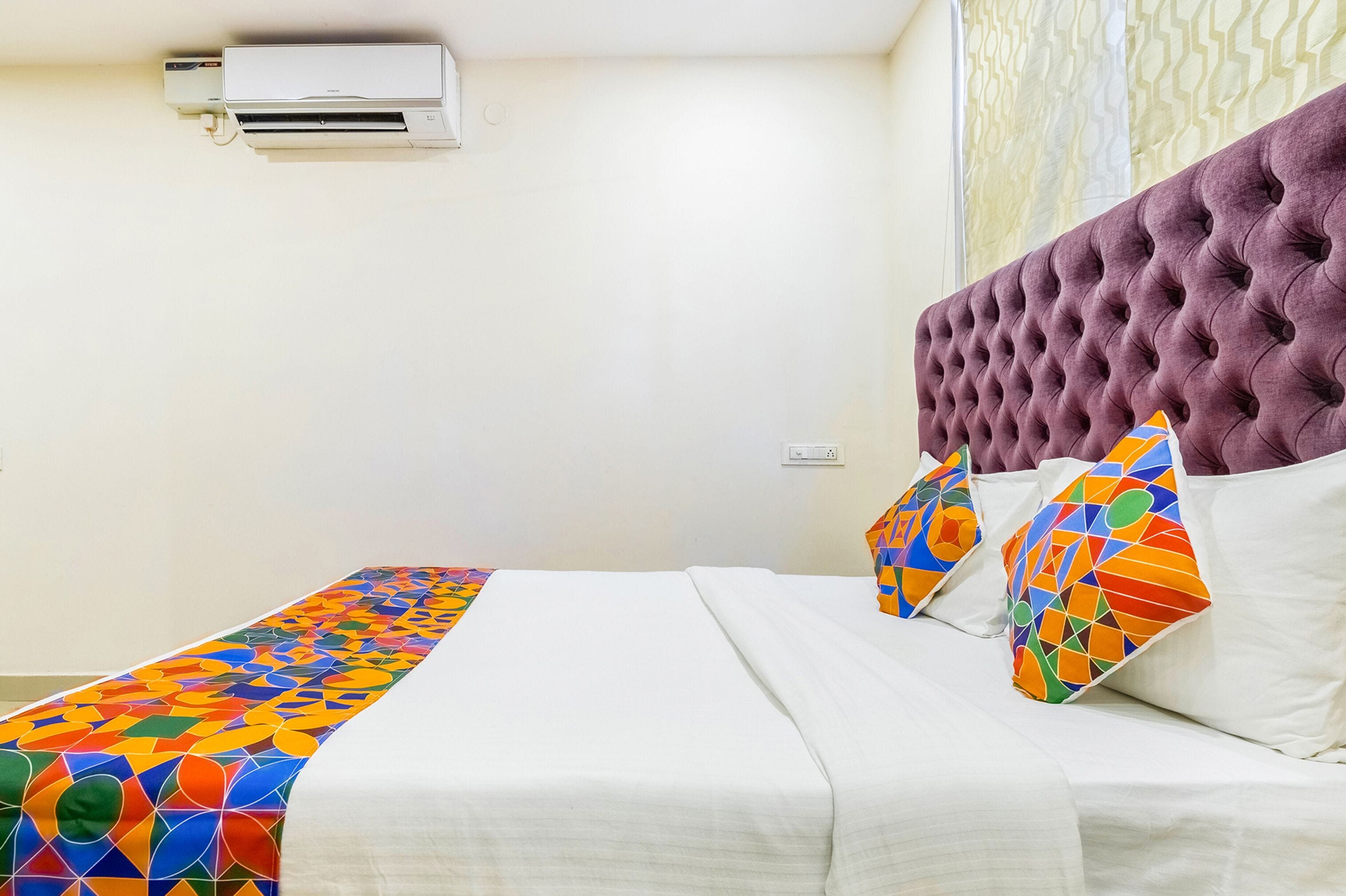 Hotelkamer met een bed, een kleurrijke, geometrisch gemasterde bedsprei, een gestuukt hoofdeinde en een airconditioningunit aan de muur.