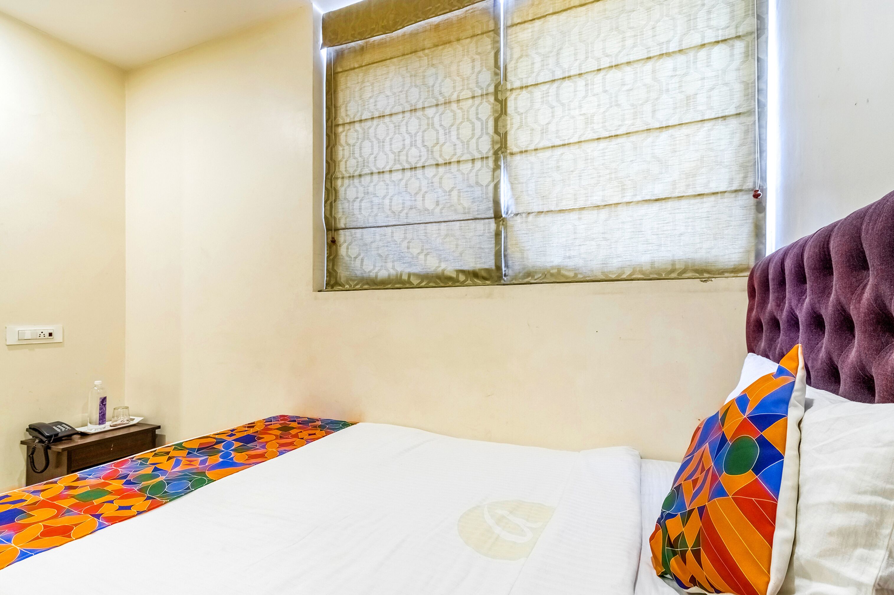 Ein Hotelzimmer mit Bett, Fenster mit gemusterten Vorhängen, Nachttisch mit Telefon und Glas, und einer farbenfrohen Tagesdecke.