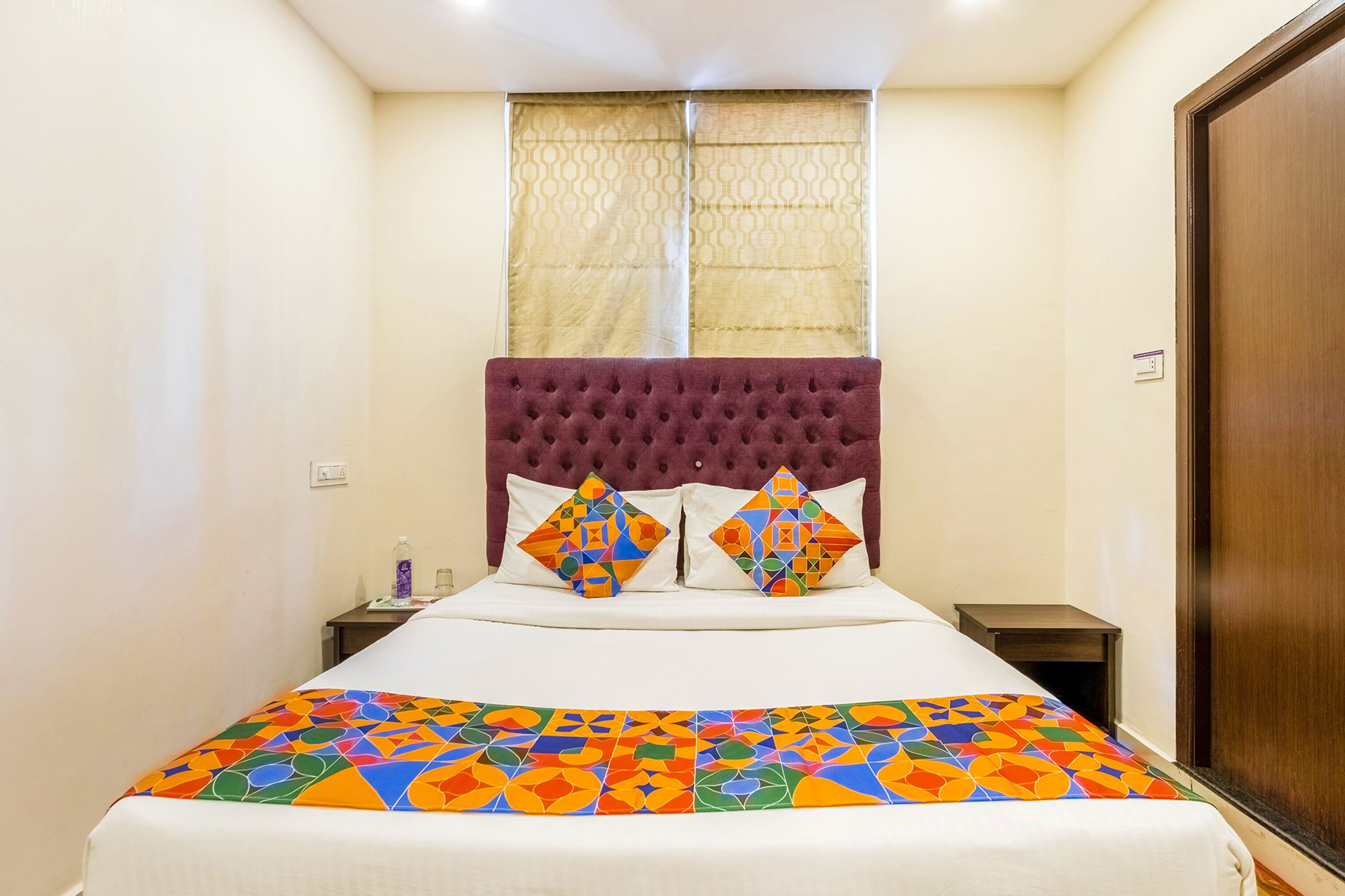A hotel room with a large bed, a tufted headboard, two bedside tables, and a window with patterned curtains.