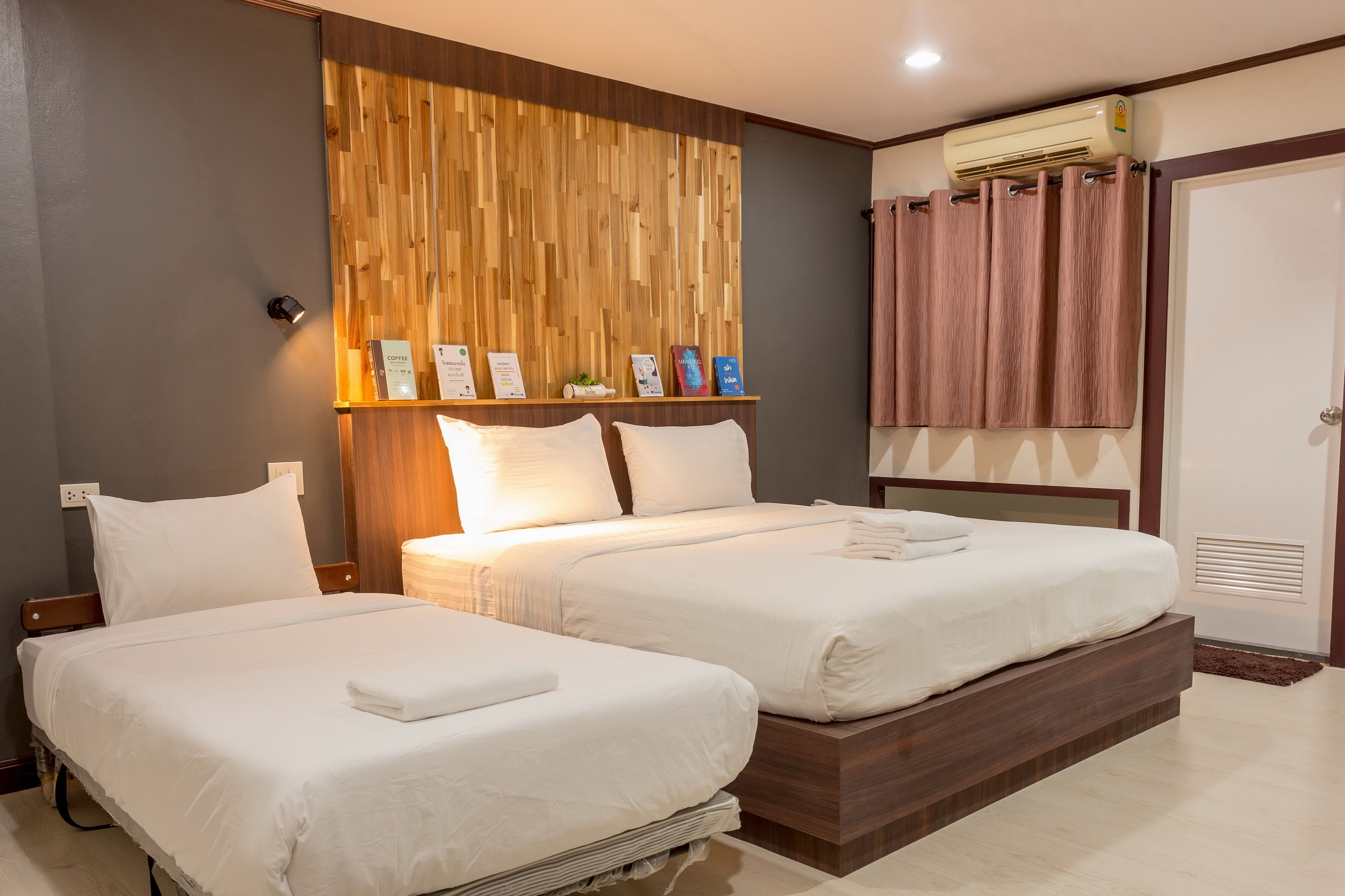 A hotel room with two beds, a wooden wall panel, a shelf with informational cards, a wall-mounted air conditioner, and a door with a curtain.