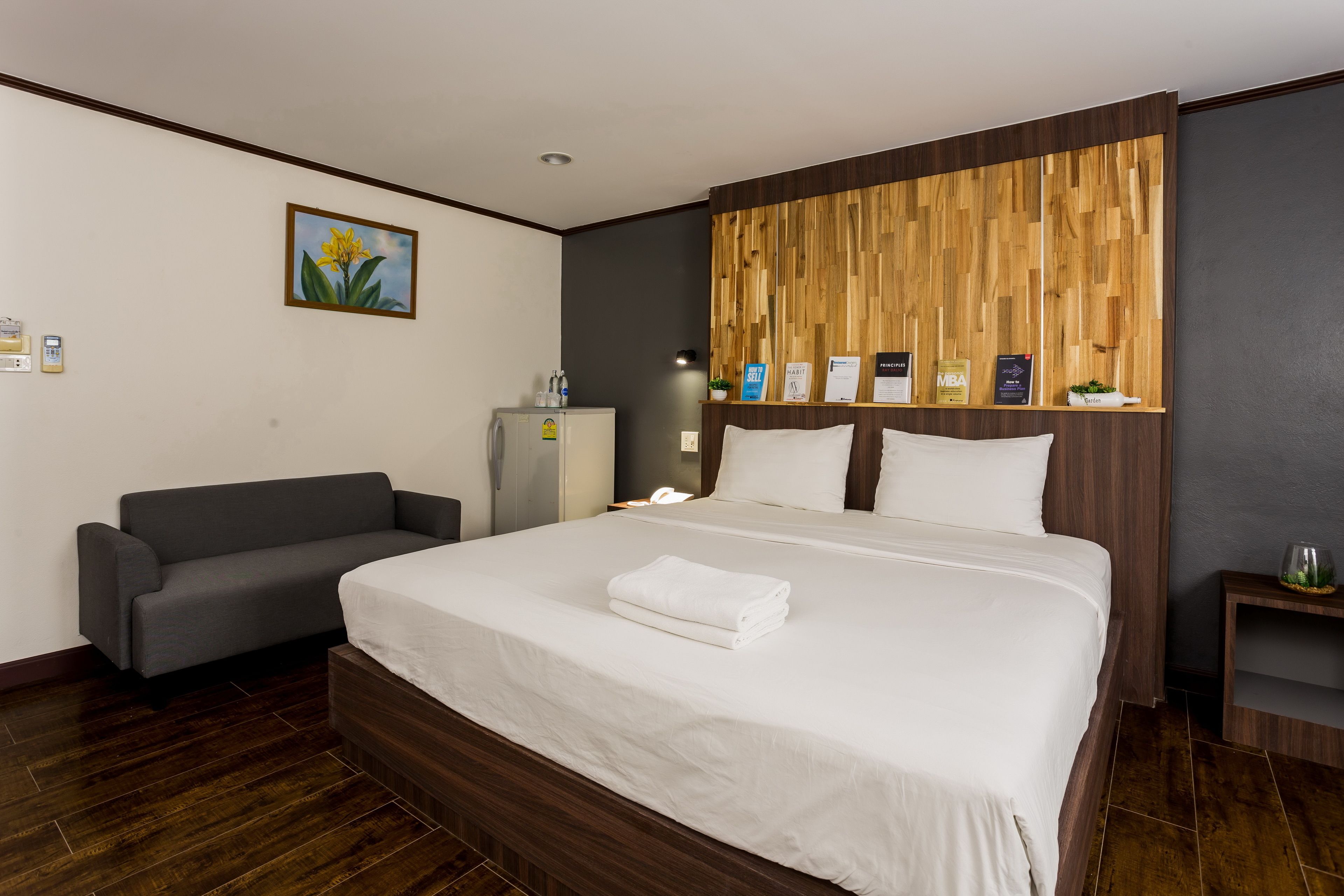 Hotelkamer met een groot bed, een grijze bank, een houten hoofdbord, een klein tafeltje met een plant en een wandplank met boeken.